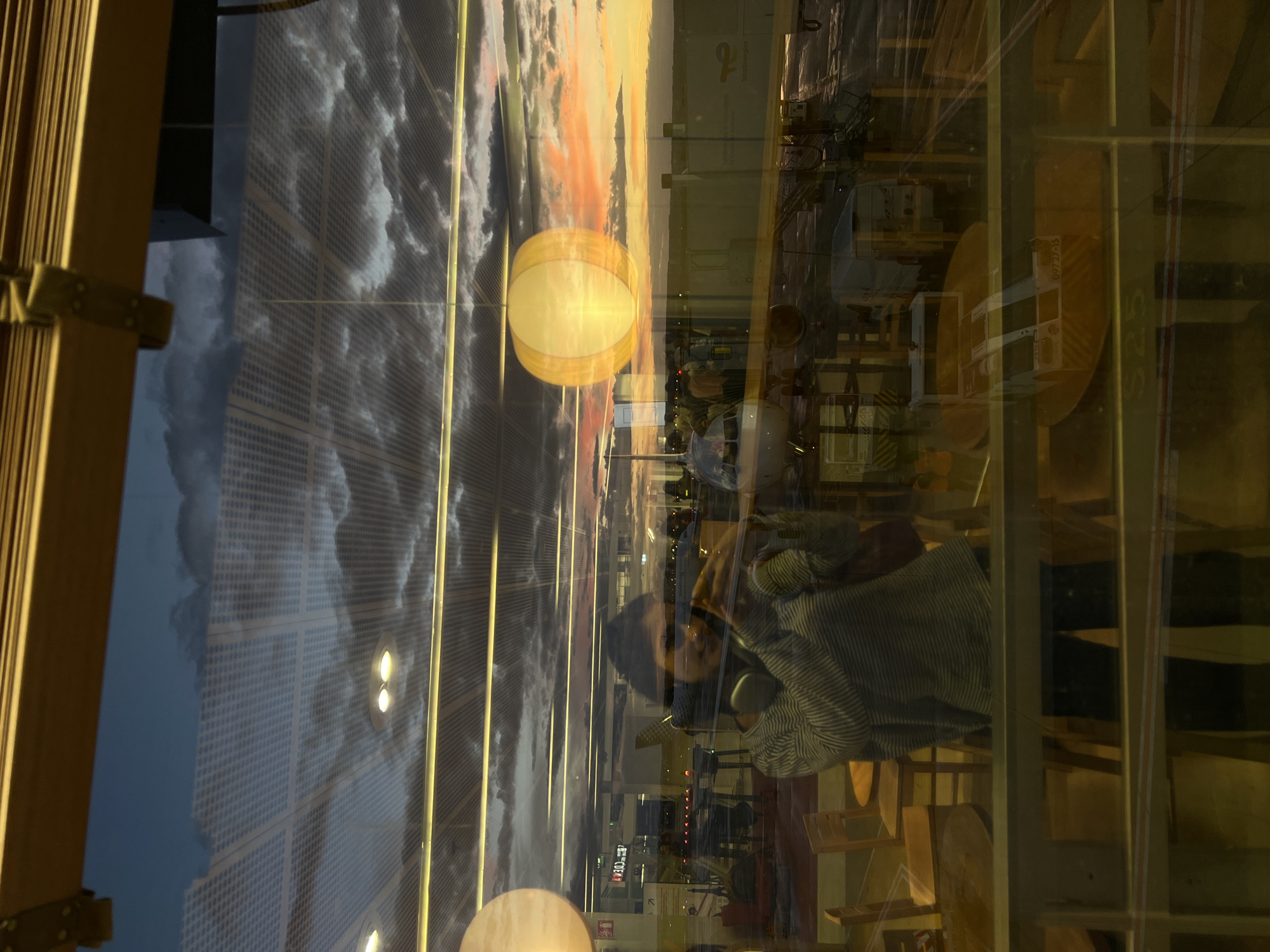 The image depicts a reflection in a window, likely at an airport, given the presence of an airplane visible through the glass. The scene is illuminated by a sunset, casting warm hues across the sky and reflecting on the glass. Inside the building, a person is visible, taking a selfie with a phone. The person is wearing headphones around their neck and a striped sweater. The interior also shows furniture, including chairs and tables. The exterior scene shows an airplane parked at a gate, with jet bridges connecting to the terminal. The overall atmosphere suggests a travel or waiting scenario during a picturesque sunset. The location is identified as Paray-Vieille-Poste, France, which is known for hosting Paris-Orly Airport.