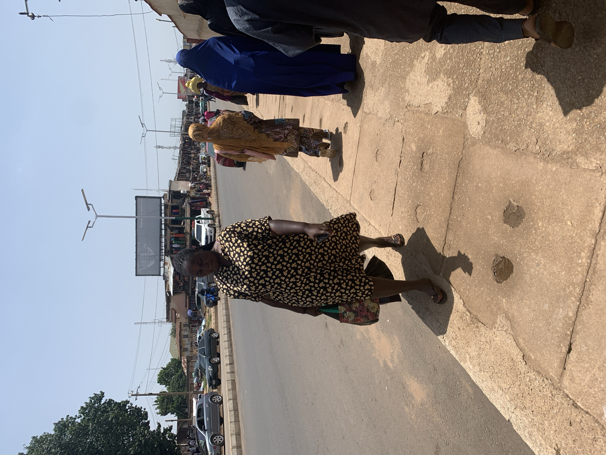 The image depicts a street scene in Jos, Nigeria. The foreground is dominated by a cracked concrete sidewalk and the asphalt road. A Black woman wearing a short-sleeved, yellow and black floral print dress and brown flip-flops walks along the sidewalk, carrying a patterned tote bag in her left hand and what appears to be a black bag in her right hand. Her shadow is cast on the sidewalk behind her.
Behind her, other individuals are walking on the sidewalk. To the right of the woman in the floral dress, a person in a vibrant blue headscarf and long blue garment is visible, followed by a person wearing a yellow headscarf and patterned clothing. Further in the background, more people are seen walking and standing.
The street is lined with buildings that appear to be shops or market stalls, with awnings and various goods displayed. Several cars, including a silver sedan and a dark green sedan, are parked or moving on the road. Traffic lights and streetlights are visible along the street, along with power lines and utility poles. The sky is clear and blue, indicating a sunny day. The overall environment suggests a busy urban commercial area.