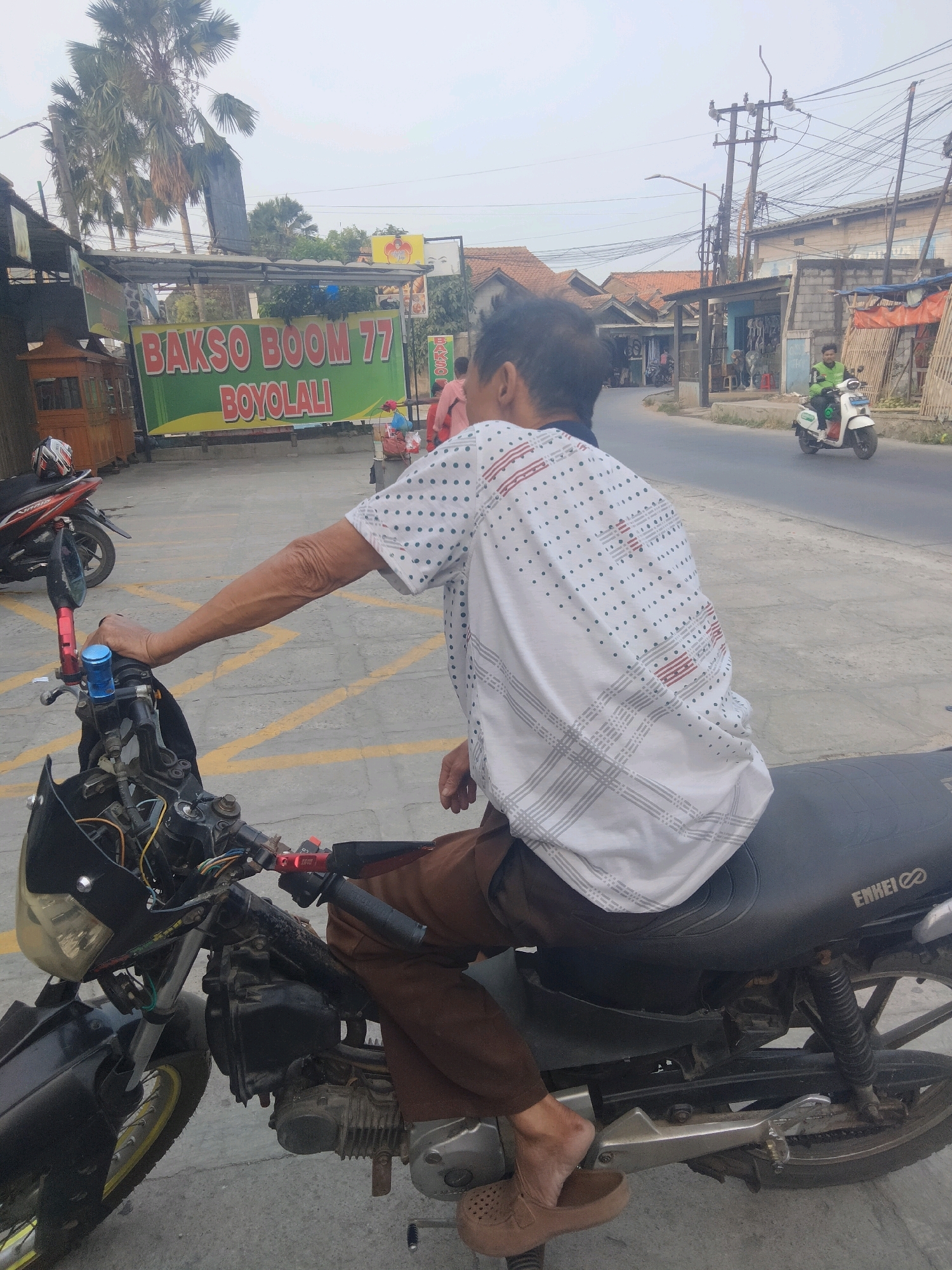 The image depicts a street scene in Pisangan Jaya, Indonesia. In the foreground, a person is seated on a motorcycle, facing away from the camera. The person is wearing a white shirt with a dotted pattern and brown pants. Their left arm is resting on the handlebars of the motorcycle, which is black and shows signs of age and use. Yellow lines are visible on the asphalt surface.
In the background, a banner with red lettering reads "BAKSO BOOM 77 BOYOLALI". Beyond that, a street is lined with various buildings, some appearing to be shops. A utility pole with numerous wires stands prominently. A white scooter with a rider in a green jacket is seen moving on the road. Palm trees are visible in the distance. The sky is overcast.