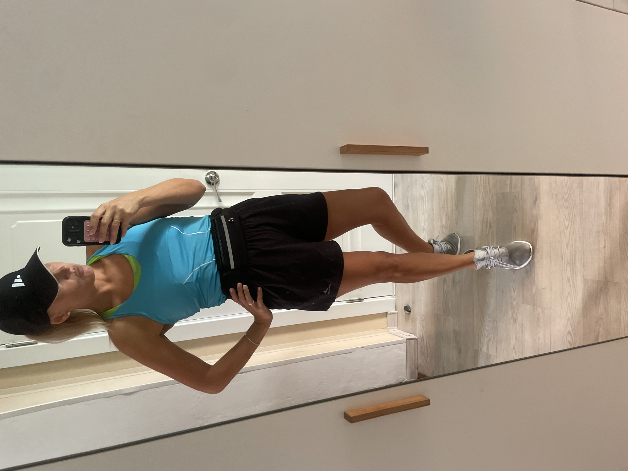A woman is standing in front of a mirror, taking a selfie. She is wearing a black baseball cap, a blue tank top with a green lining, black athletic shorts, and grey sneakers. She has a black fanny pack around her waist and is wearing a bracelet and a ring. The environment appears to be a room with light-colored walls and a wooden floor. There is a closet door with a wooden handle to her right.