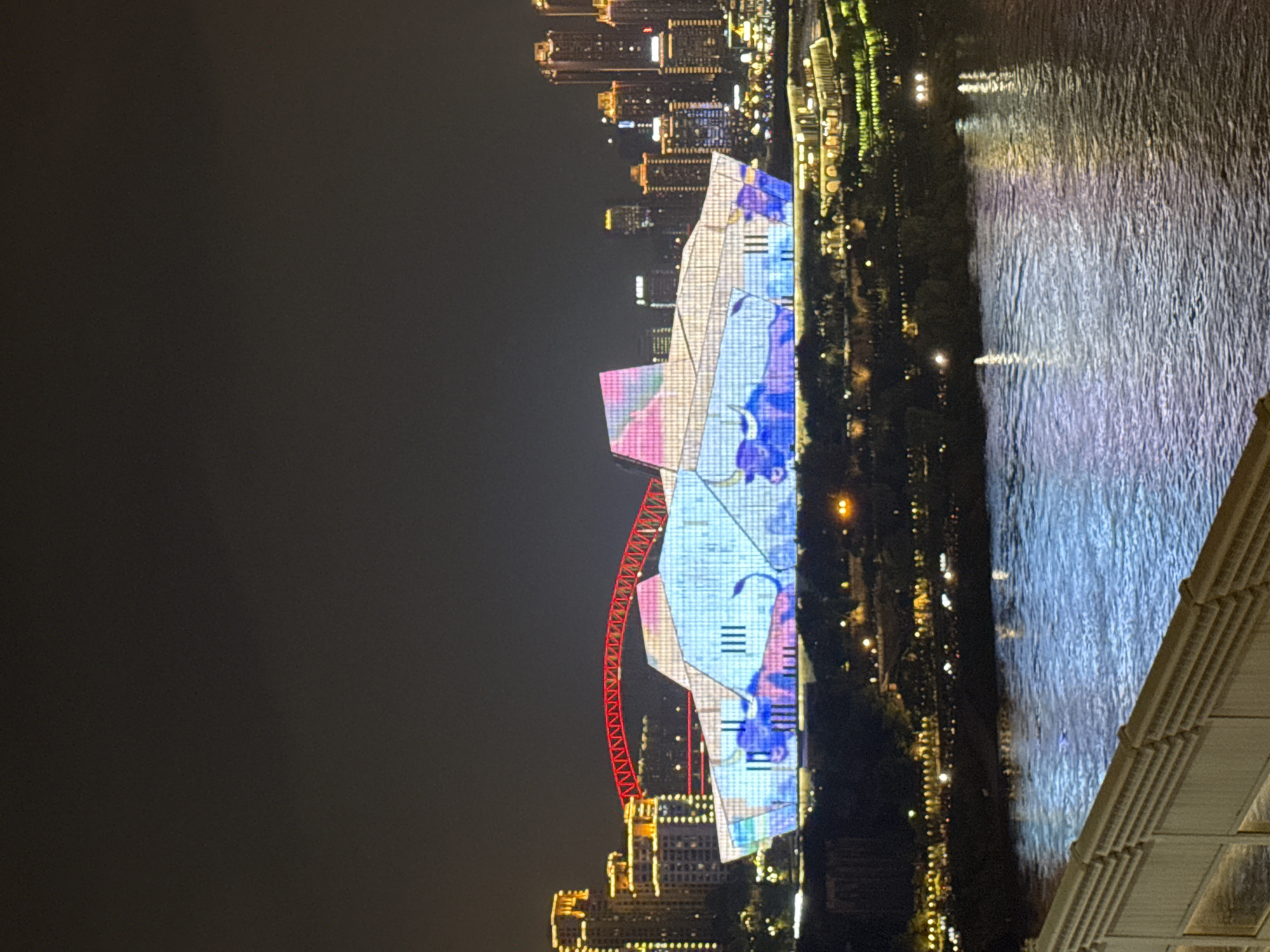 An eye-level, wide shot captures the illuminated facade of a large, modern building at night in Yuzhong District, China. The building's geometric, angular design is covered in a light display featuring blue and pink abstract patterns and depictions of bulls. A prominent red, arched bridge spans the sky behind the building, with a city skyline of numerous illuminated skyscrapers in the background. The foreground shows a body of water reflecting the city lights, and a railing of a bridge or elevated walkway is visible in the bottom left corner. The overall scene is one of urban nighttime splendor and architectural dynamism.