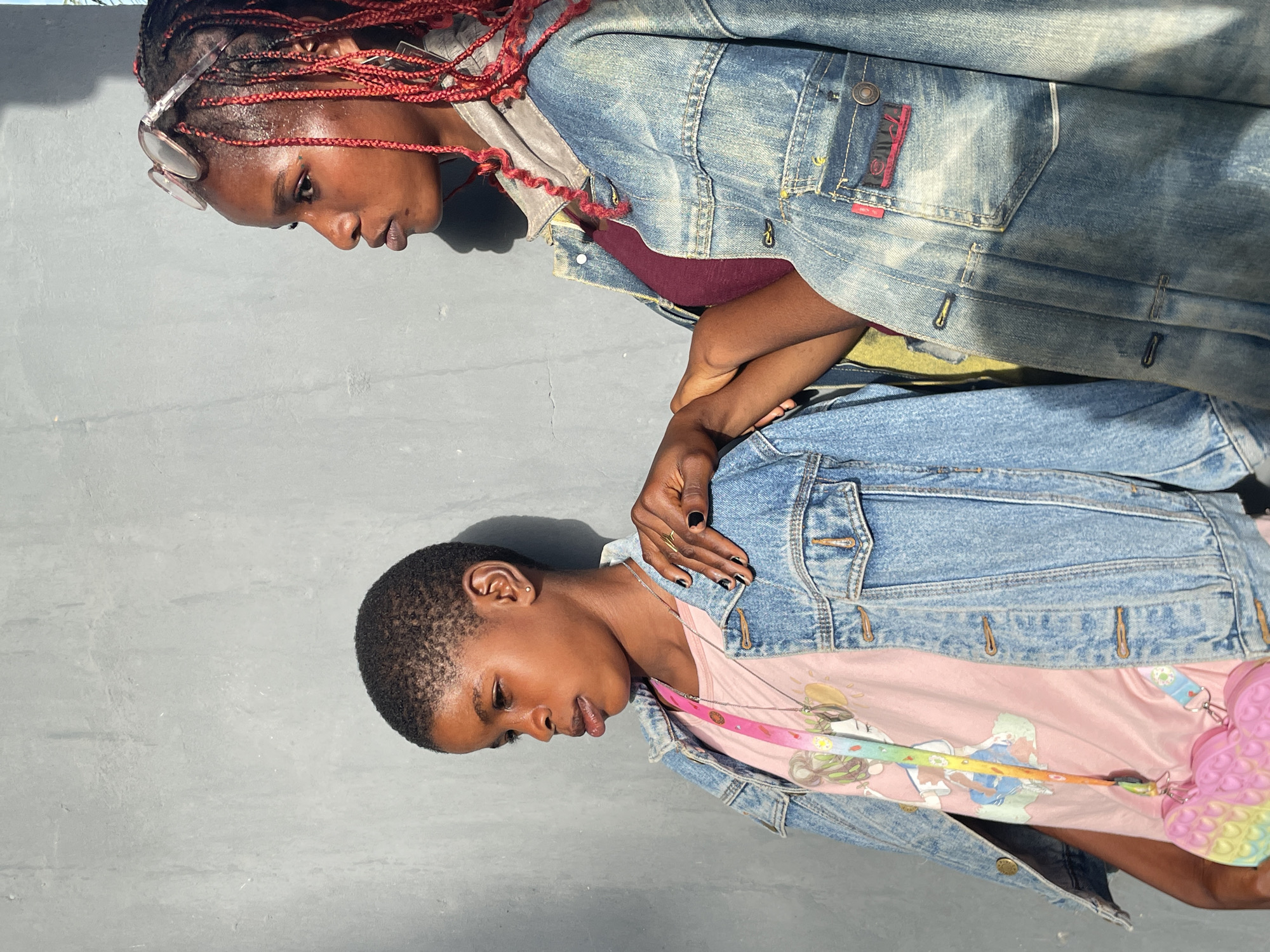 Two dark-skinned individuals, likely young women, are captured in a portrait against a plain, light gray concrete wall in Badagry, Nigeria. Bright daylight illuminates the scene, casting clear shadows, suggesting a sunny daytime setting.
The person on the left has long, red braided hair and wears light-framed sunglasses pushed up on their head. They are dressed in a light blue denim jacket over a dark red top, with their gaze directed downwards. On the pocket of their denim jacket, a small red label with faint white text, possibly a brand name, is visible.
The person on the right has short, closely-cropped dark hair. They wear a light blue denim jacket over a pink t-shirt featuring a colorful, cartoon-like graphic design. Their left hand, adorned with black painted fingernails and a small ring, gently rests on the arm of the person beside them. A vibrant, rainbow-colored lanyard with a butterfly-shaped 'pop-it' toy hangs from their neck. Their expression is calm and contemplative, with eyes cast downwards.
The individuals appear to be posing together for a photograph, displaying a relaxed and reflective mood.