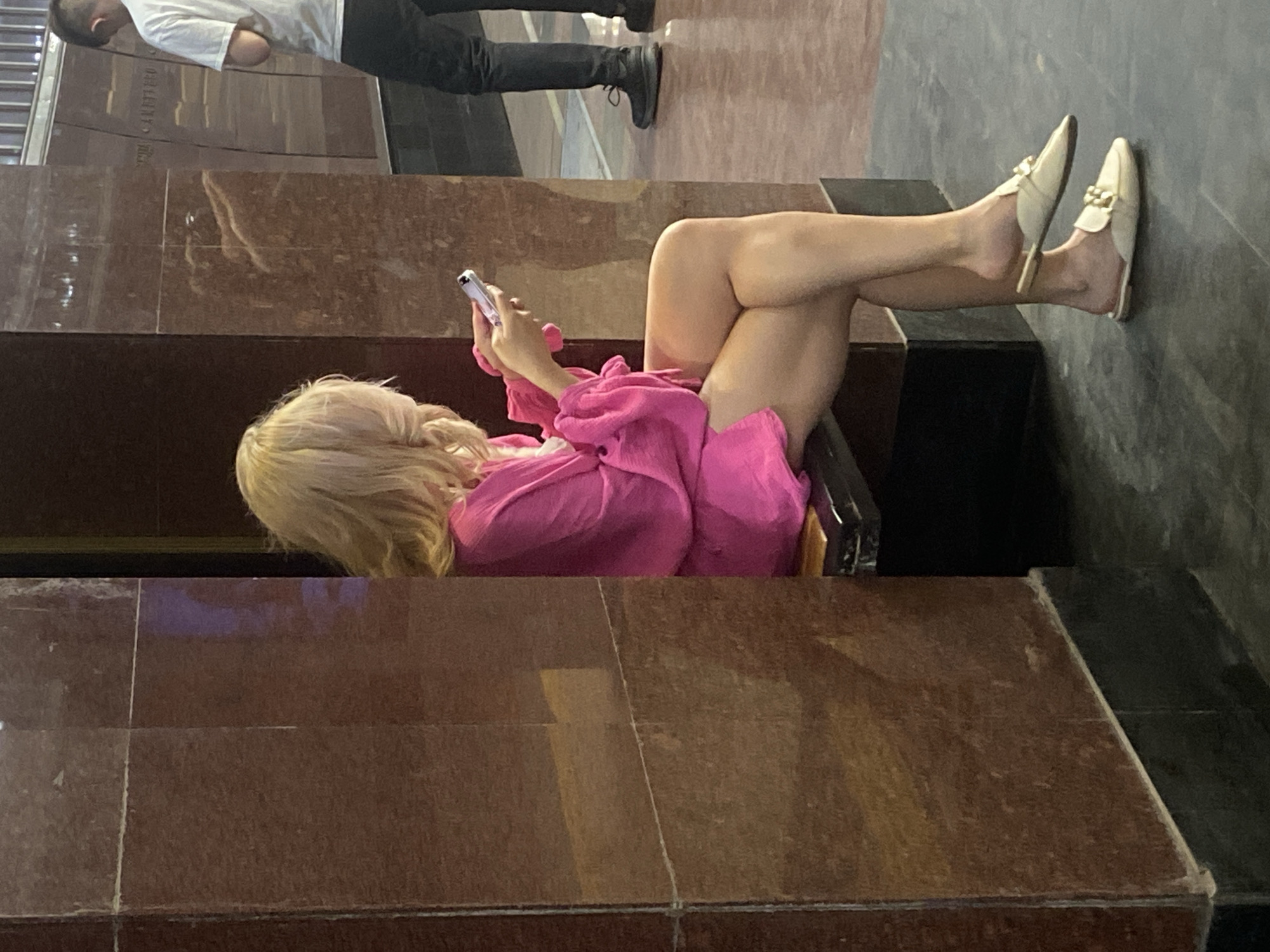 The image depicts a person lying on a ledge with legs extended, while using a smartphone. The individual has blonde hair and is wearing a pink outfit. The person is wearing beige or light colored loafers. Another person in a white shirt is partially visible above on a different ledge. The surrounding environment features a granite or marble-like material with a tiled or segmented pattern. Given the architectural style and the provided information, this image is likely taken in Kazan, Russia.