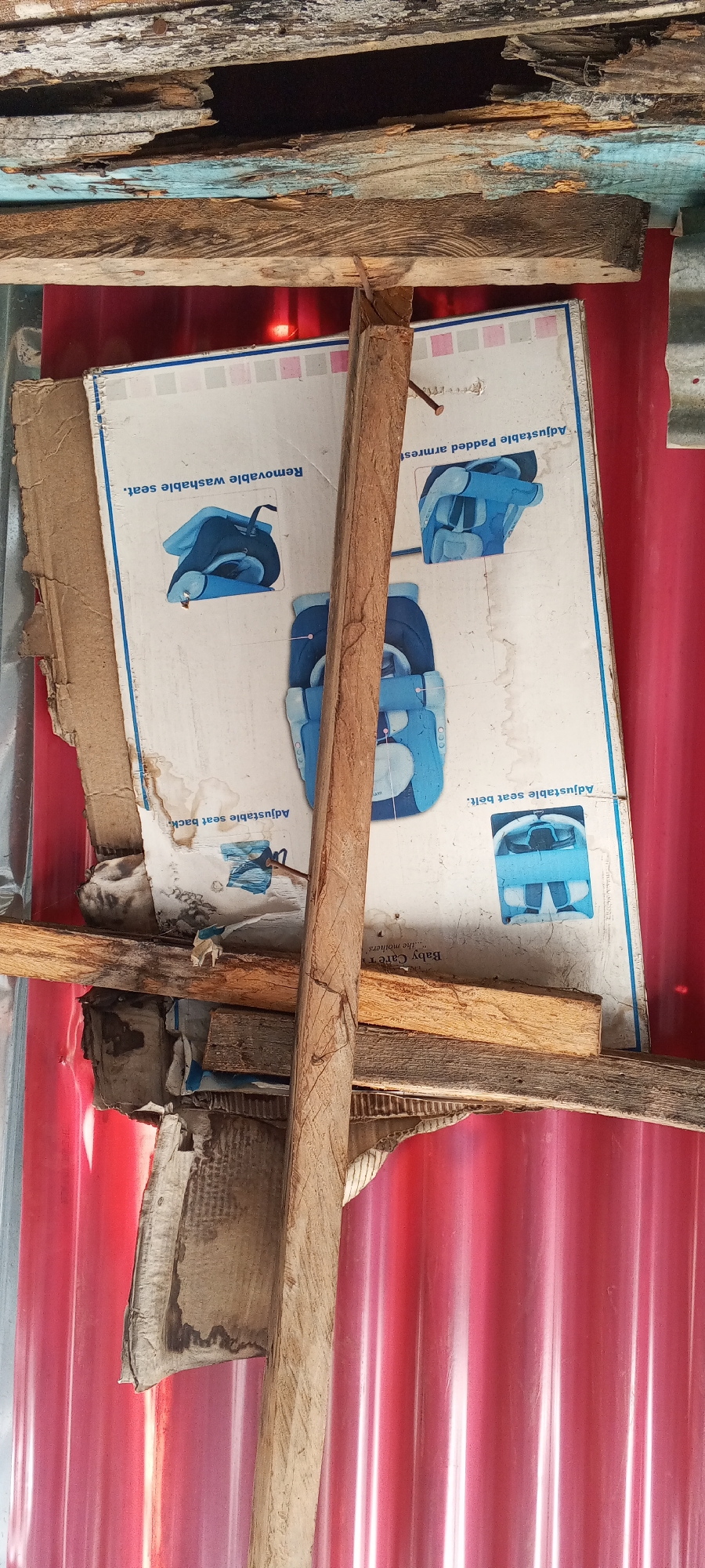 The image depicts a close-up view of a damaged cardboard box, likely from a baby car seat, propped up against a corrugated red metal wall. The box displays illustrations of a blue car seat, with labels such as "Removable washable seat" and "Adjustable Padded armrest." It is supported by several rough wooden planks and appears to be in a state of disrepair, with torn edges and water stains. The overall environment suggests an outdoor or makeshift structure, possibly a storage area or a temporary shelter. The background is dominated by the red corrugated metal, and the top of the image shows weathered wooden beams. The scene is consistent with a location in Jalingo, Nigeria, where such materials and building styles are common in informal settlements or construction sites. There are no people or active interactions visible in the image.