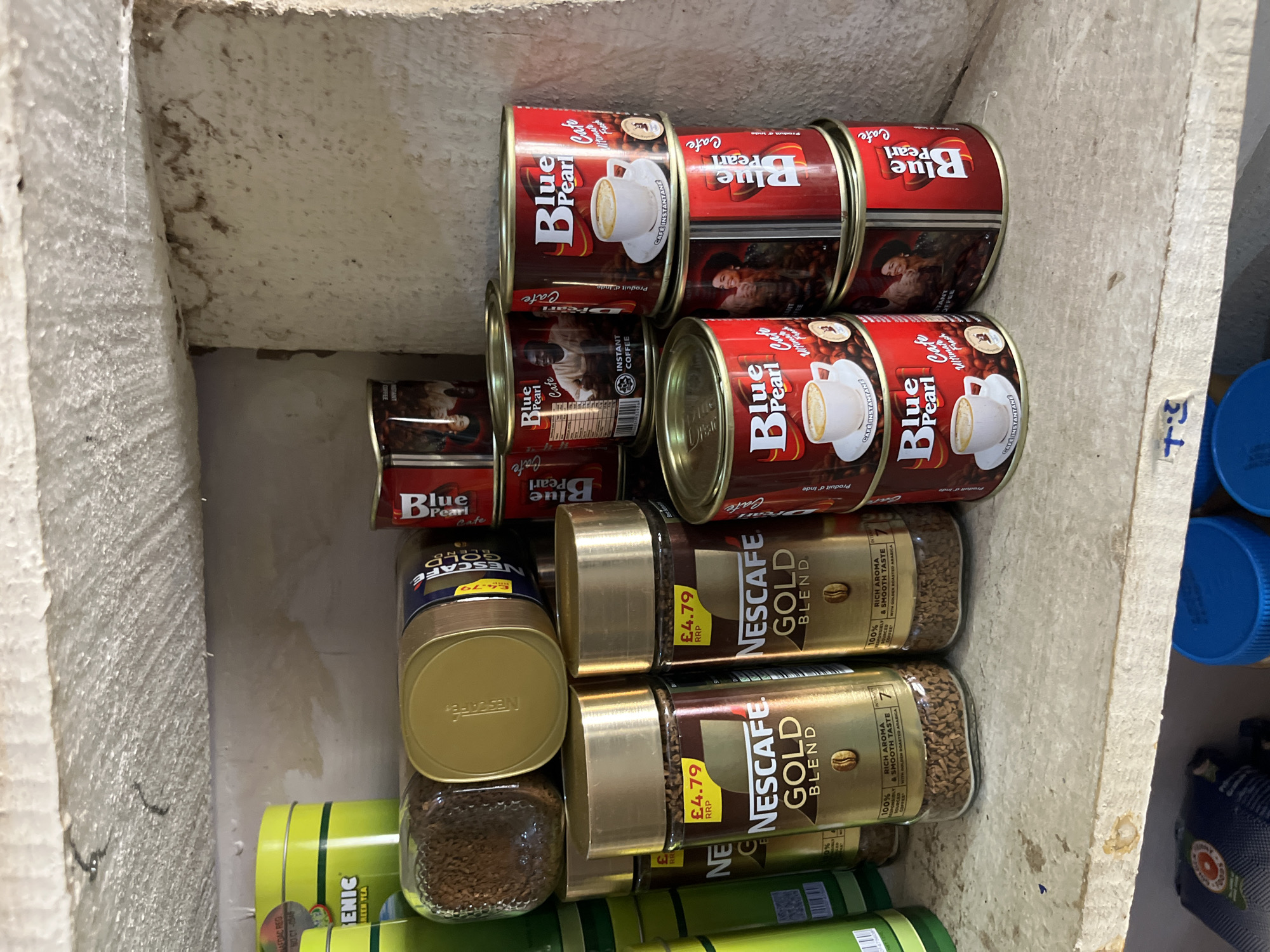 A low-angle, full shot captures a shelf in a storage area in Bukuru, Nigeria, filled with various brands of coffee and tea products. The shelf itself is rough and textured, showing signs of wear and tear with some staining. In the foreground, two large glass jars of Nescafé Gold Blend coffee sit side-by-side, each featuring a £4.79 RRP price tag and a gold lid. Behind them, a larger, wider jar of Nescafé Gold Blend coffee is positioned on its side, also with a gold lid. To the left of the Nescafé jars, part of a bright yellow cylindrical tin labeled "ENIC GREEN TEA" is visible.
Dominating the right side of the image are stacked red cylindrical cans of "Blue Pearl Cafe" instant coffee. Several cans are visible, some stacked two high, and others forming a partial third layer. These cans feature images of coffee cups and people enjoying coffee. Some of the cans have text indicating "INSTANT COFFEE" and "Produit d'Inde" (Product of India). The overall impression is one of a retail or storage space stocked with coffee and tea products, likely in a market or small shop setting.
