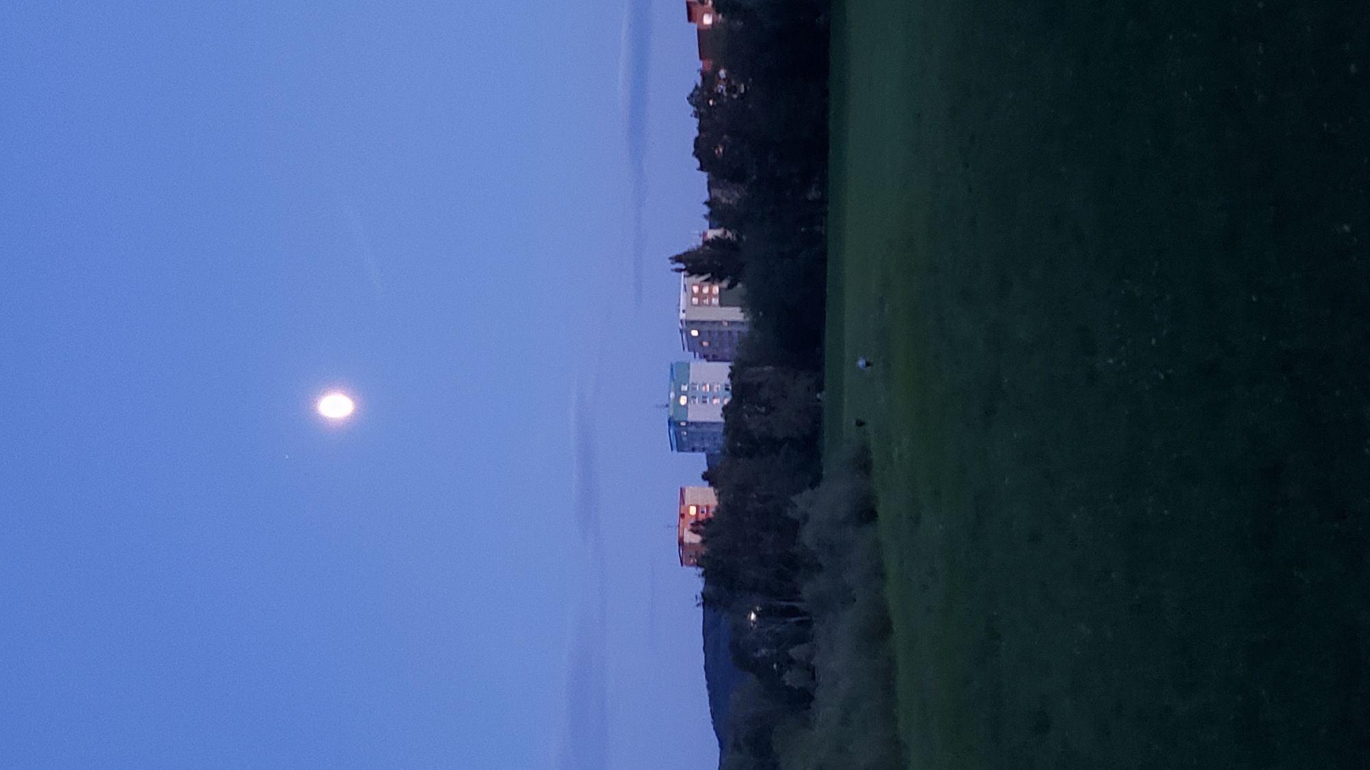 An outdoor, low-angle, vertical medium shot captures a dusky twilight scene over a field and residential buildings in Prešov, Slovakia. The sky is a deep, clear blue with a bright, full moon positioned in the upper center, accompanied by a smaller, fainter celestial body to its left. In the mid-ground, a line of trees separates the open field from a row of apartment buildings. The buildings, illuminated with internal lights, appear to be of medium height, with varying color facades including orange, white, and blue. The foreground consists of a dark, green, grassy field, with a few small, dark figures of people scattered across it, appearing to be walking. The overall atmosphere is serene and twilight.