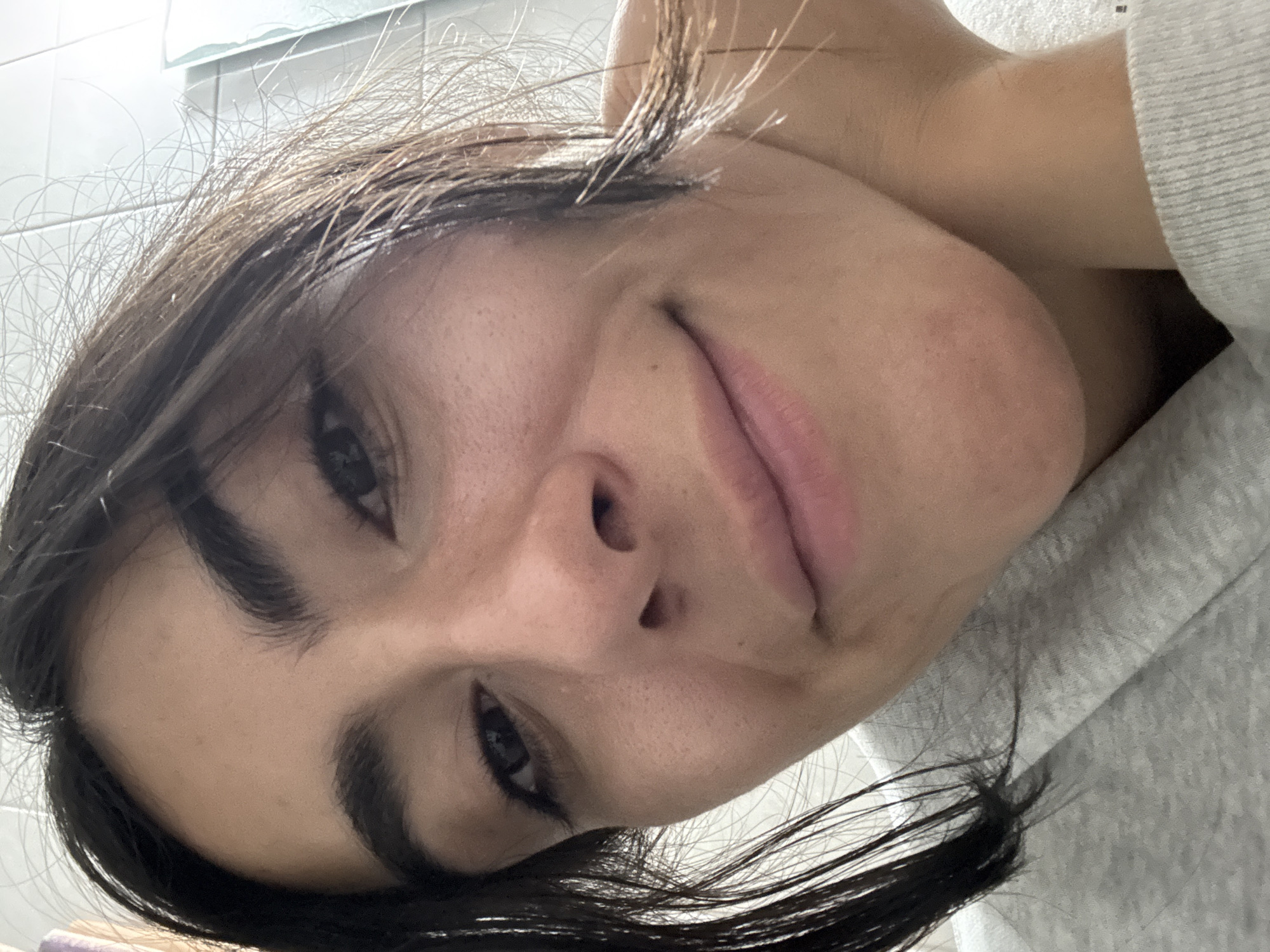 A close-up selfie of a young woman with dark hair and light skin. She is wearing a light gray sweatshirt. The background appears to be a tiled bathroom wall. No specific details indicate the location is Puebla, Mexico.