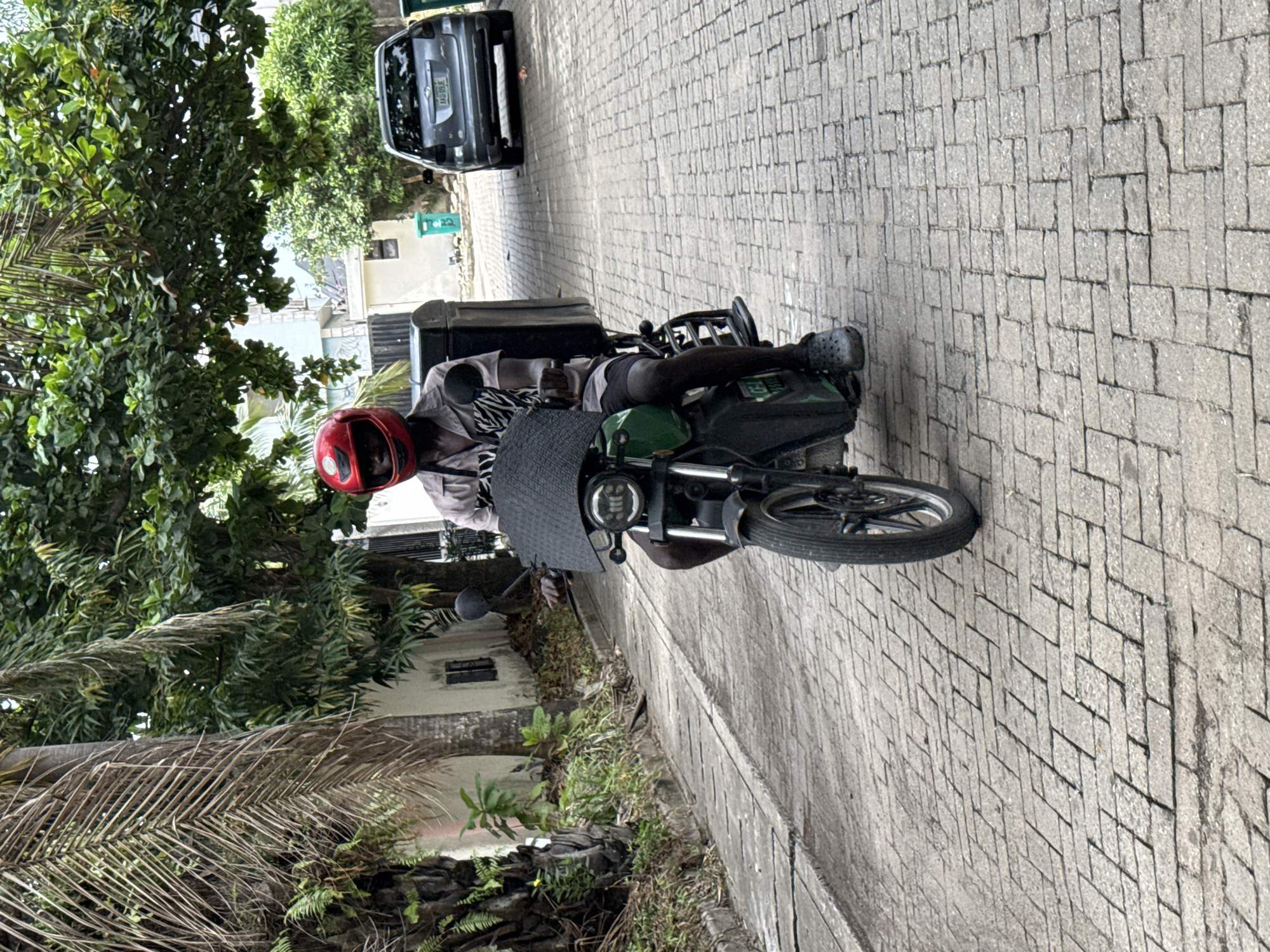 A daytime image captures a person on a motorcycle parked on a grey paved road composed of interlocking bricks. The person, positioned facing towards the right side of the frame in the reoriented view, wears a red helmet, a light-colored shirt over a zebra-striped long-sleeve top, dark shorts, and grey Crocs. They are seated on a green motorcycle that features a black cargo box mounted behind the seat and a textured dark grey windshield.
Lush green foliage, including dense trees and palm fronds (some dried), dominates the left side of the frame, indicating a tropical or subtropical environment. In the background, further down the paved road, a dark grey or black sedan is parked. Its license plate is partially visible and reads "MOWSE". A small green utility box with the number "22" on its side can also be seen near the car.
The scene depicts a quiet street or paved area, likely in Lekki, Nigeria, given the context. The bright, even lighting suggests a clear, sunny day. The person on the motorcycle appears to be stationary, perhaps waiting or paused before continuing their journey. No discernible emotions are visible due to the helmet obscuring the face.