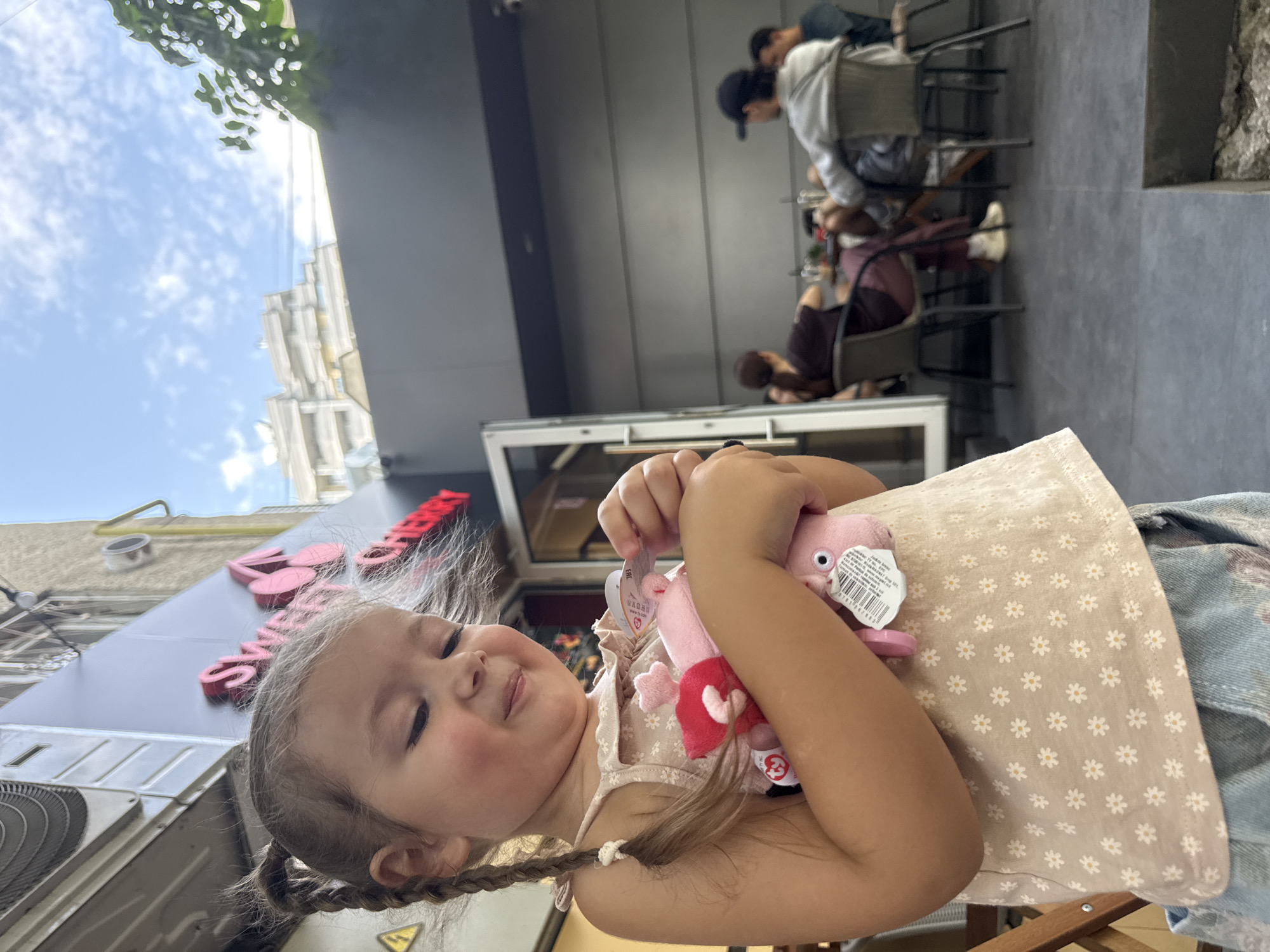 In Chișinău, Moldova, a young girl with braided hair holds two pink stuffed animals, one with red accents, close to her chest. She wears a light-colored dress with a floral pattern and denim shorts. Her eyes are closed and she has a gentle smile. She stands in front of a building with a sign that reads "SWEET CHERRY" in large, pink, stylized letters. The background shows an outdoor seating area of a cafe with several people seated at tables. The sky above is blue with scattered white clouds.