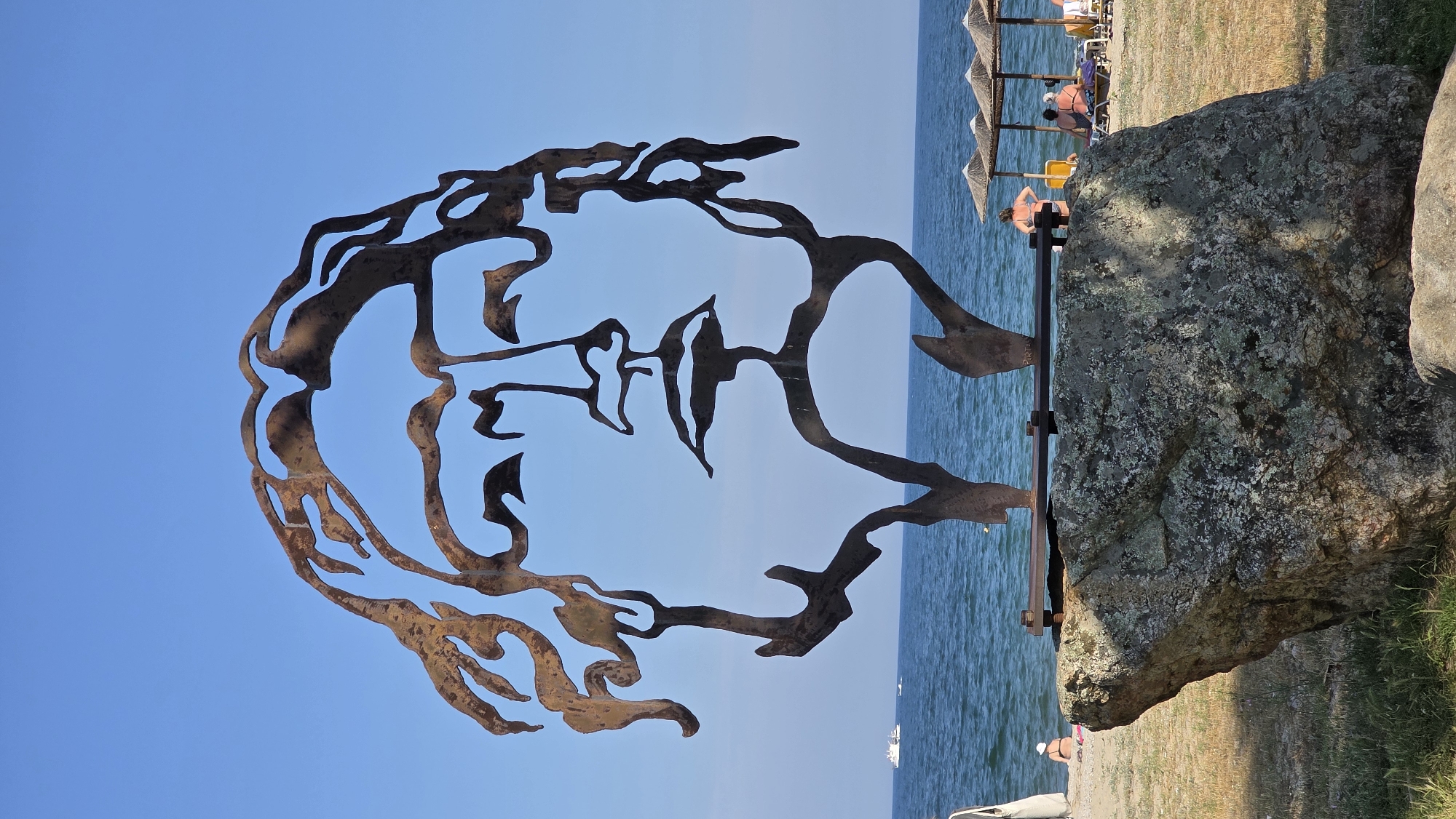 The image displays a large, rusted metal sculpture of a stylized human face, centrally positioned on a beach in Asprovalta, Greece. The sculpture, characterized by an outline form with defined curly hair, eyes, nose, and full lips, is mounted on a substantial, irregularly shaped rock base. The rock's surface exhibits a textured appearance with visible lichen or moss. The sculpture is attached to the rock by a horizontal metal bar secured with bolts.
In the background, a calm, blue sea extends beneath a clear, bright blue sky. On the left side of the sea, a white motor yacht and at least two smaller vessels are visible. The sandy beach is visible between the sea and the foreground rock. Several individuals are present on the beach. On the left, a person in a bikini is seated near the water's edge, partially obscured by the rock base. Further left, part of a person carrying a light-colored bag is visible. On the right side of the beach, multiple conical straw umbrellas are erected, shading sun loungers occupied by bathers. One person in a bikini stands facing the sea near these umbrellas. The immediate foreground around the rock base consists of dry grass and sand. The scene is brightly illuminated by direct sunlight.