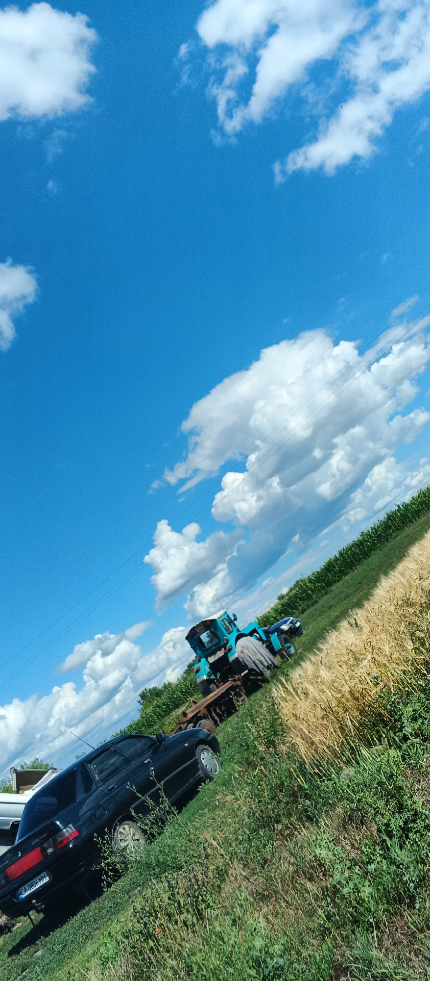 The image, captured in Ternivka, Ukraine, depicts an outdoor scene under a bright blue sky with scattered white clouds. In the foreground, a black sedan is parked on a grassy slope, its rear facing the viewer. The car has a visible license plate that reads "CA 0806 MH". Behind the sedan, a blue tractor is positioned on a field. The tractor appears to be hitched to a piece of farming equipment, possibly a plow. The field itself is a mix of green vegetation and golden crops, suggesting a rural or agricultural setting. The scene is characterized by natural lighting and a slightly tilted perspective, possibly due to the way the photo was taken. There are no people visible in the image.