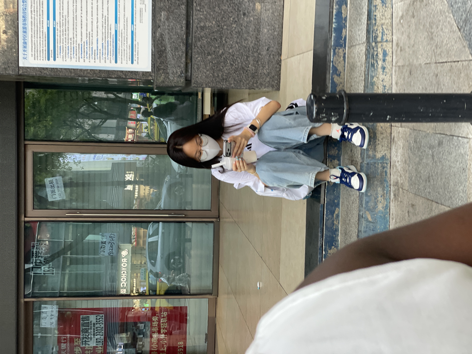 A young East Asian woman wearing a white t-shirt, blue jeans, and a face mask sits on stone steps outside a building with large glass doors. She is holding a phone and a beverage cup with a straw. The building appears to be a commercial establishment, with signs in Chinese visible through the glass. In the background, vehicles and pedestrians are present on a street. The scene is located in the Yuzhong District of China.
