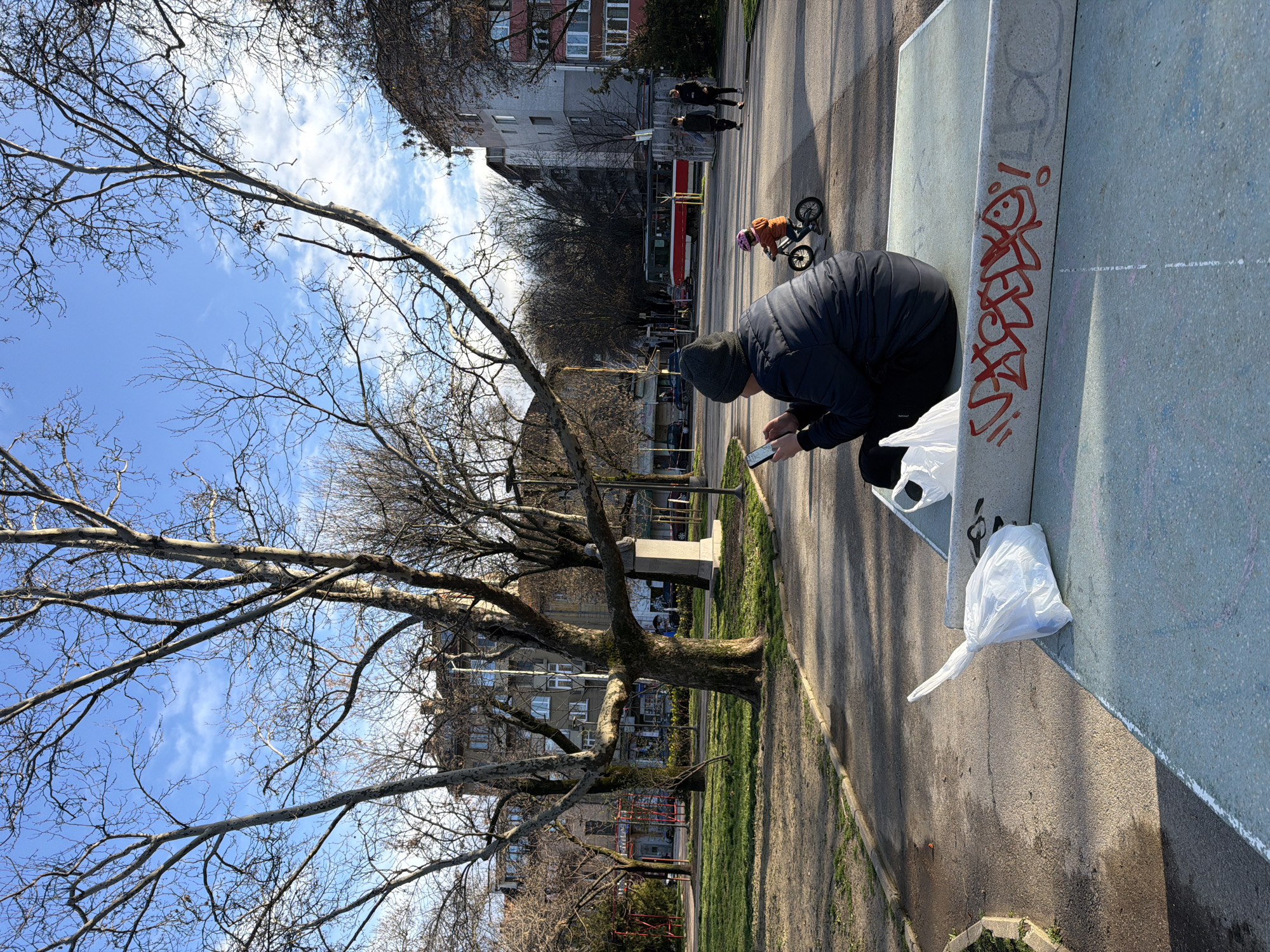 The image portrays an outdoor urban park scene within Doctor's Garden (Doktorska Gradina) in Sofia, Bulgaria, under a clear blue sky with scattered white clouds. In the immediate foreground, a person is seated with their back to the viewer on a light blue-gray concrete surface, its vertical face marked with red graffiti reading "VXOX!" accompanied by a smiley face. This individual wears a dark blue puffer jacket and a dark gray beanie, holding and looking at a white smartphone. Two white plastic bags are positioned on the concrete surface to the left of the seated person. Paved asphalt pathways traverse the park, flanked by grassy areas. A small child, wearing an orange jacket and a pink helmet, rides a black balance bike on a paved path to the right of the foreground figure. Further along the path, two adult figures walk away from the camera. Bare deciduous trees with light-colored bark, notably a large plane tree in the midground, are prominent. To the left, a red metal playground structure is partially visible, and to the right of the large tree stands a stone monument topped with a bust. In the background, a street features parked cars and a red and white trolleybus. Beyond the street, multi-story residential buildings with varying architectural styles, characteristic of Sofia, are visible. The scene is illuminated by strong natural sunlight, casting visible shadows.