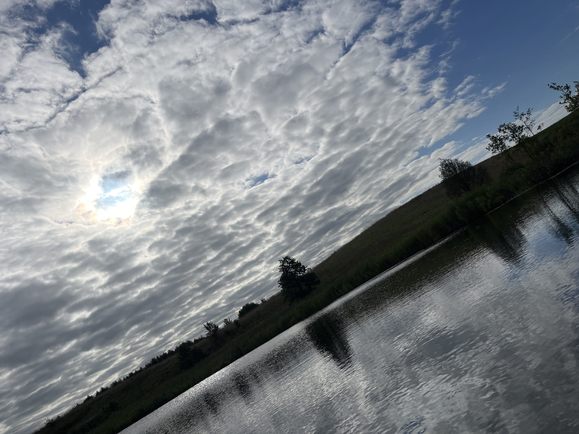 The image shows a diagonal view of a body of water reflecting the sky and a grassy embankment with scattered trees. The sky is filled with rippling, fragmented clouds, with a bright spot indicating the sun peeking through. The embankment, covered in green grass and some darker vegetation, slopes down towards the water. The water itself is dark and reflects the patterns of the clouds and sky. The overall scene suggests an outdoor, natural environment, possibly a lake or pond, located in the rural landscape of Дунілавіцкі сельскі Савет, Belarus. There are no people or animals visible in the image, and no specific actions or interactions are depicted. The lighting indicates it is daytime, with the sun partially obscured.