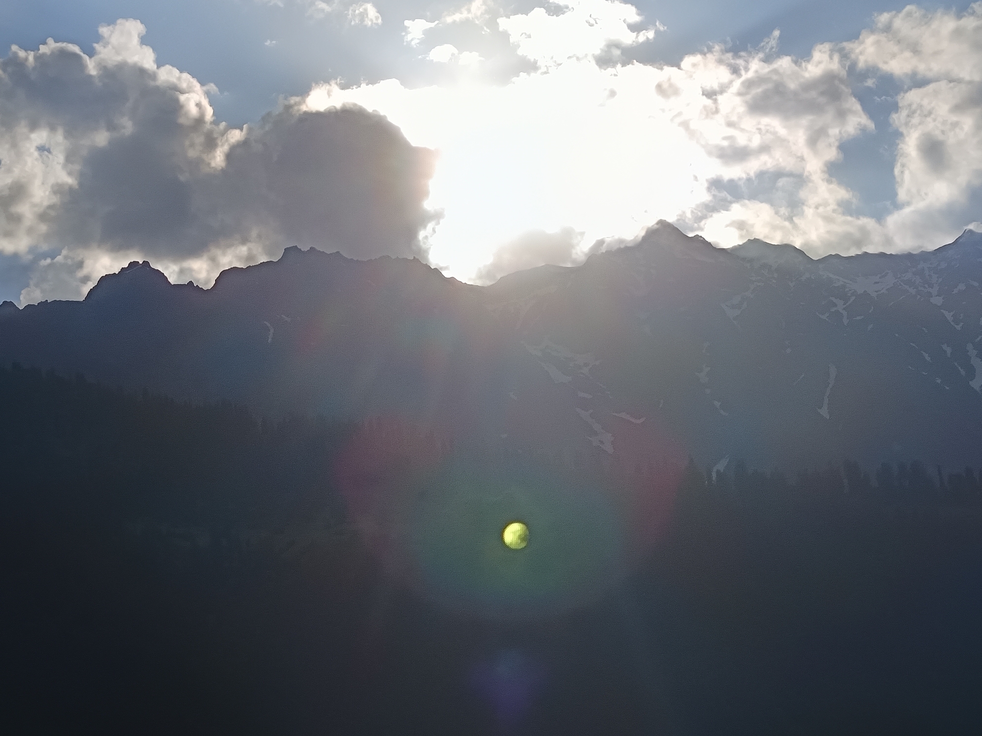The image displays a mountainous landscape under a bright, partly cloudy sky, characteristic of Vashisht, India. The sun is positioned behind a large bank of clouds in the upper-middle, generating a powerful backlight that silhouettes the terrain and creates significant lens flare. The upper portion of the sky is blue with scattered white and grey clouds, some illuminated from behind. A prominent mountain range occupies the middle ground, its rugged peaks appearing dark against the bright sky, with visible patches of snow on the slopes towards the right. The lower half of the image is composed of extremely dark, indistinct terrain, likely forested hillsides, further obscured by heavy shadow and strong light refraction. A distinct, bright yellow-green circular lens flare is centered in the lower-middle section, accompanied by diffuse purple and red flares extending from the bright light source. No people, animals, specific man-made objects, actions, or interactions are discernible within the frame. The scene is dominated by natural elements and atmospheric lighting effects.