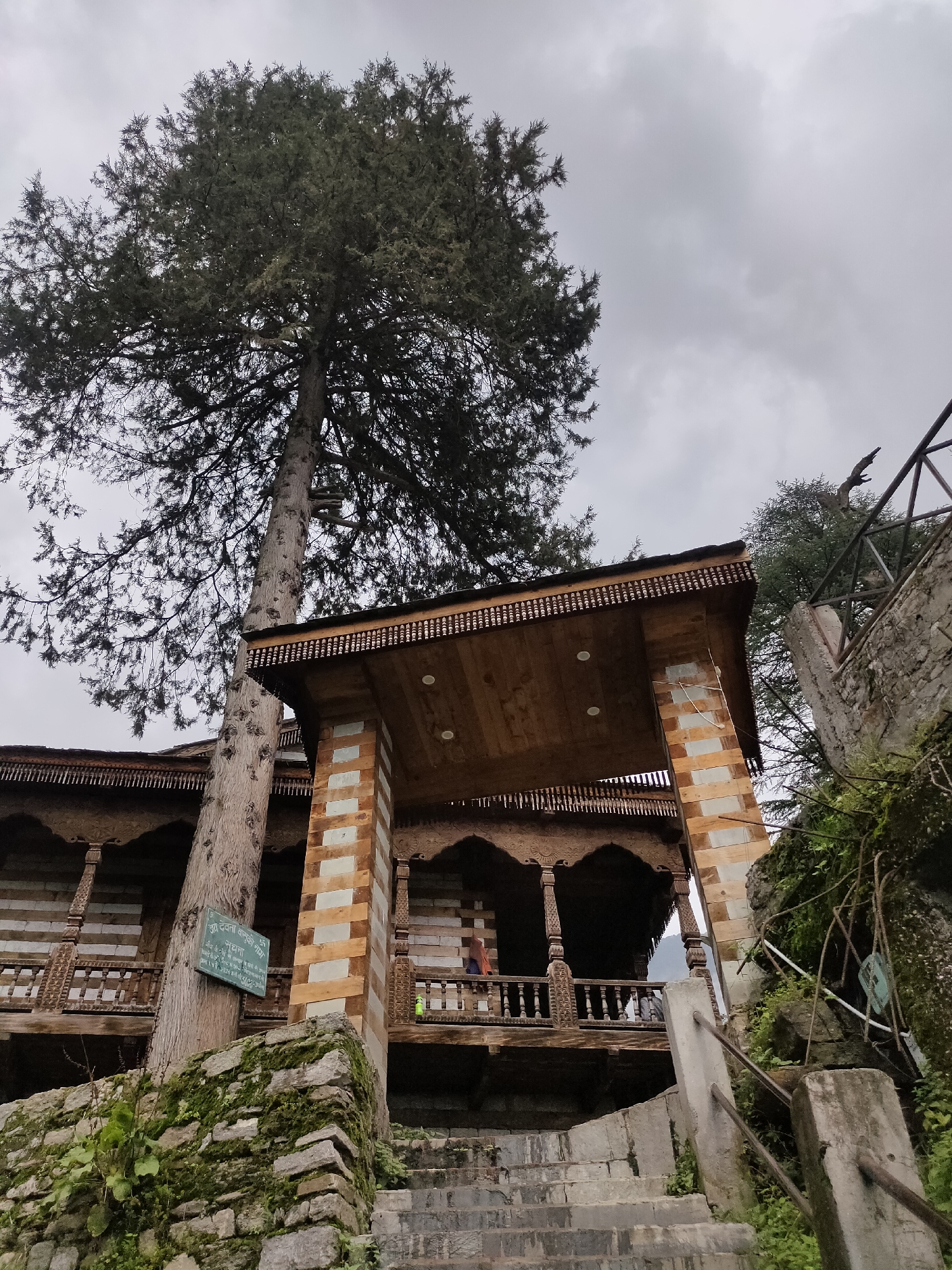 In Raman, India, a traditional wooden structure with intricate carvings is featured prominently in the image. The building, likely a temple or a heritage site, showcases detailed woodwork on its balconies and eaves. A large, mature tree stands beside the structure, its branches reaching towards a cloudy sky. Stone steps lead up to the building, with moss-covered stone walls lining the pathway. A green sign with text in an Indian script is attached to the tree. The overall impression is one of historical architecture set within a natural, somewhat overcast environment.