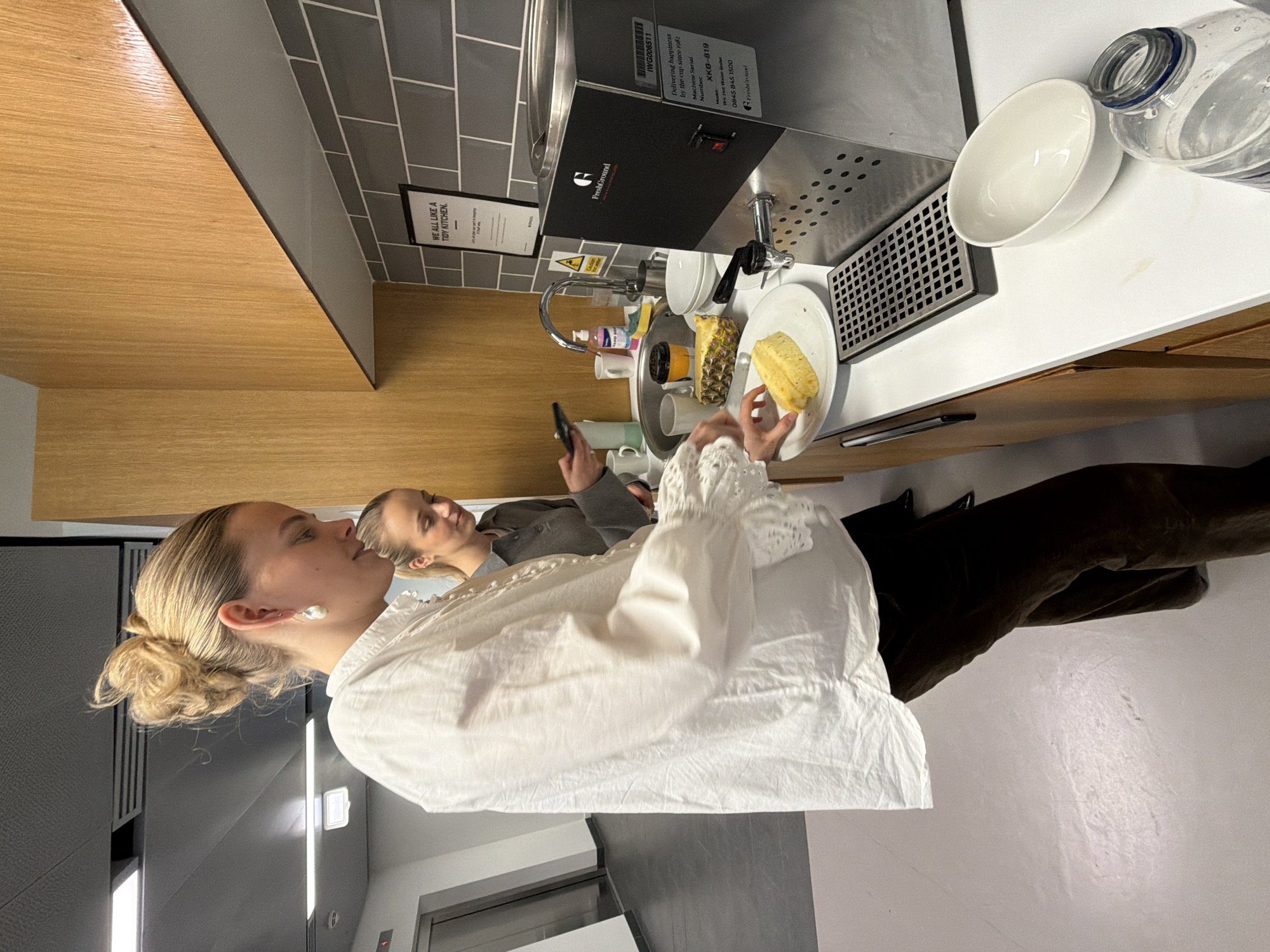 Here is a detailed description of the image:
**Scene:** The image is taken indoors, likely in a kitchen or kitchenette area of a workplace. The location is identifiable in the city of London, United Kingdom due to the presence of a hot water dispenser bearing a UK phone number with the 0845 area code, which is associated with non-geographic numbers used in the UK.
**People:** There are two women visible. One woman is closer to the camera, wearing a white blouse with lace detailing. Her blonde hair is styled in a bun. The second woman is in the background, holding a phone and wearing a grey top. Both women appear to be of Caucasian ethnicity.
**Objects:**
* Hot water dispenser with the brand name "FreshGround" visible.
* A white bowl and a water bottle on the countertop.
* A plate with slices of pineapple.
* A sink with various items, including cups and cleaning solutions.
* Tiles on the backsplash.
* Wooden cabinets.
* A sign that says "WE ALL LIKE A TIDY KITCHEN."
**Actions:**
* The woman in the foreground appears to be interacting with the pineapple.
* The woman in the background is looking at her phone.
**Interactions:** There is no evident direct interaction between the two women.
**Environment:** The room seems well-lit and clean. The decor is modern, with a combination of wooden and grey tones. The presence of the hot water dispenser and kitchen items suggests a communal area designed for preparing food and beverages.