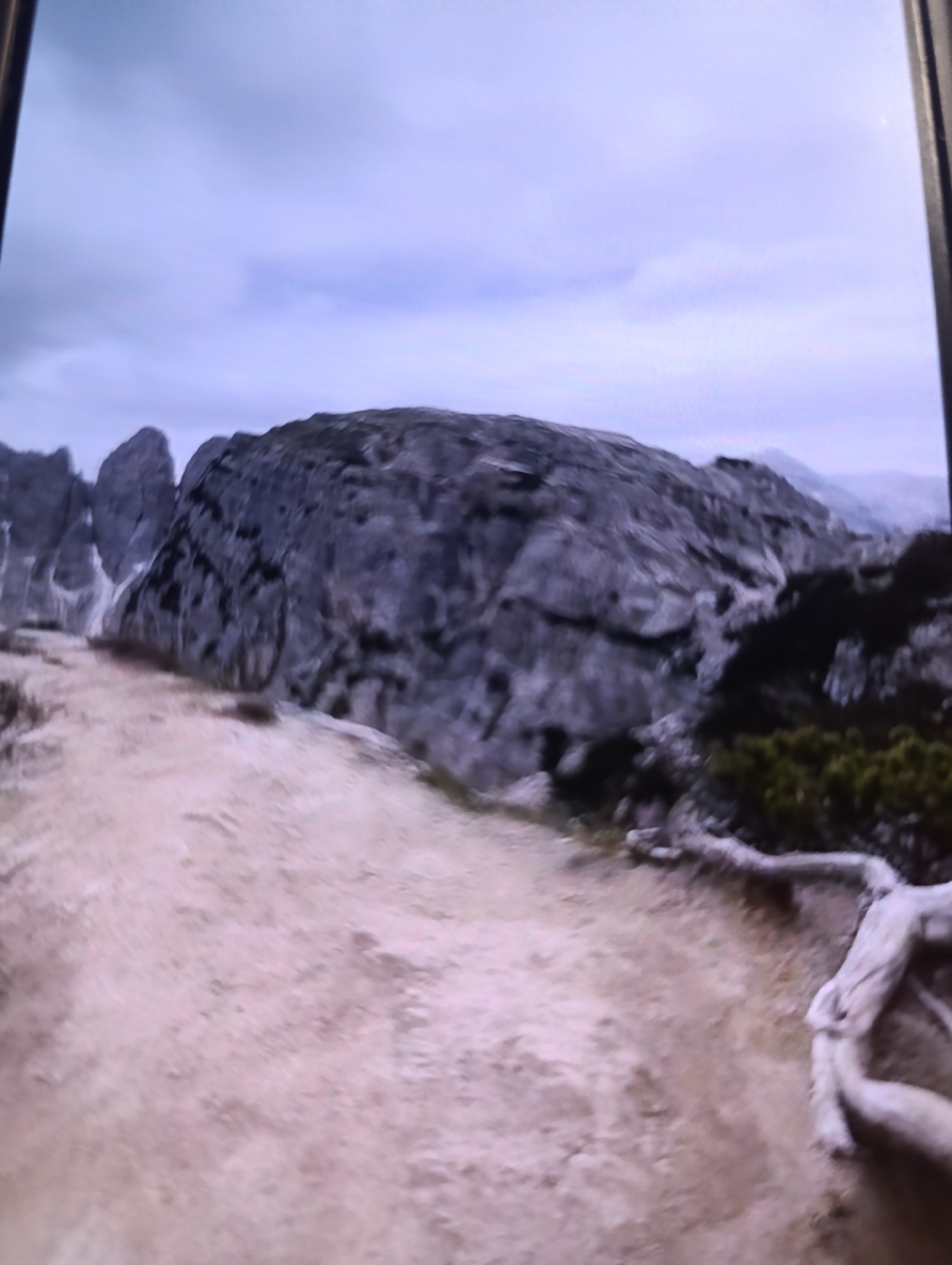The image depicts a panoramic view of a mountainous landscape under an overcast sky. The foreground is dominated by a sandy, rough terrain that leads towards a large, imposing rock formation in the midground. This rock is characterized by its rugged texture and dark coloration, with visible crevices and variations in shade. To the left of the main rock, a series of other rock formations rise, some appearing sharper and more jagged, with patches of snow or light-colored mineral deposits visible on their surfaces. The background reveals more distant mountains, shrouded in a soft haze, suggesting depth and vastness. On the right side of the midground, a patch of dark green vegetation is present, with gnarled tree roots or branches exposed on the sandy ground. The overall impression is one of a natural, untamed environment, possibly a hiking trail or a remote wilderness area.