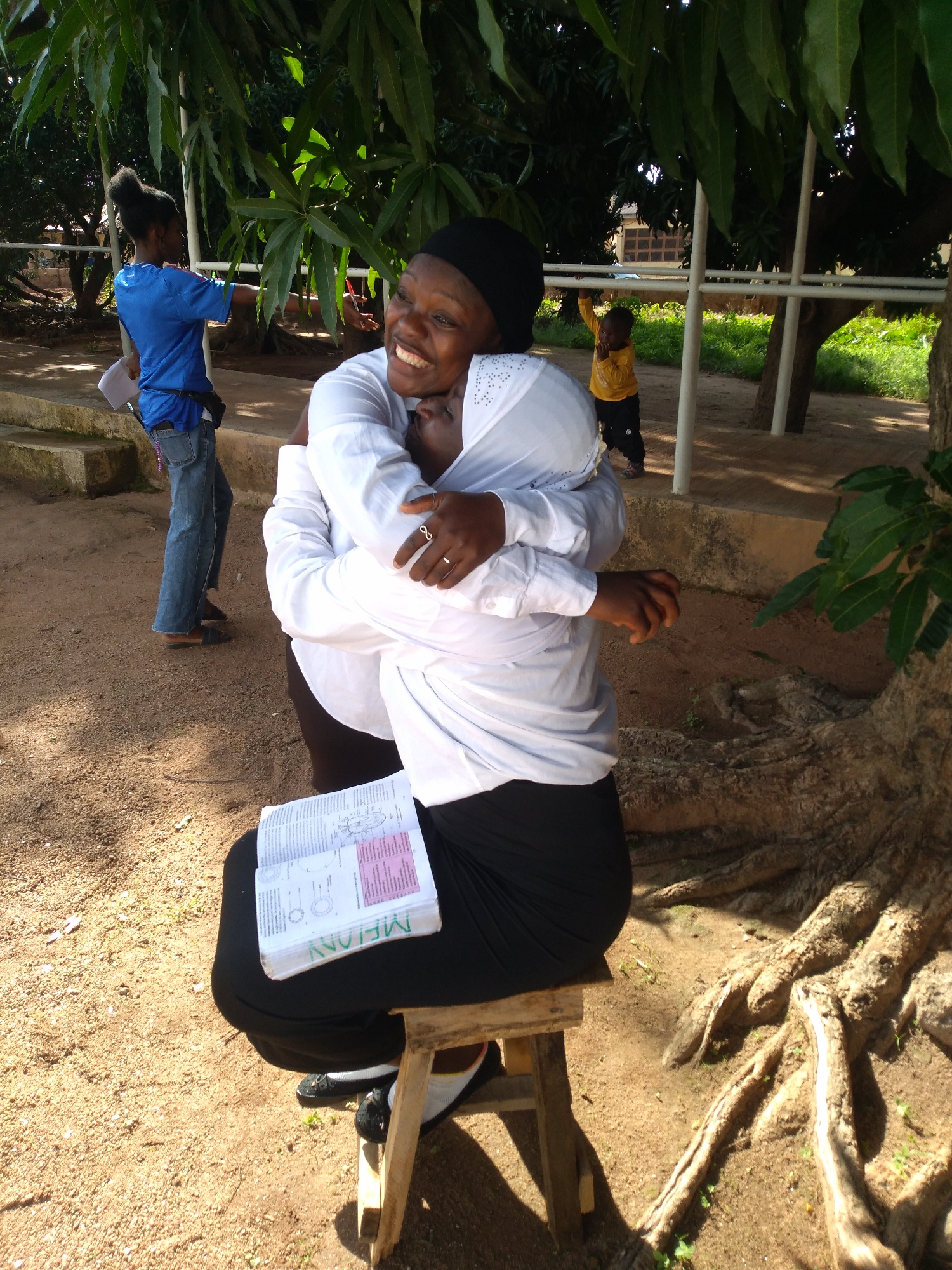 This image captures an outdoor scene in Jos, Nigeria. Two adult females are central to the composition, engaged in an embrace beneath a tree with dense foliage providing shade. One female, positioned with her face towards the camera, exhibits a wide smile. She wears a black head covering, a white long-sleeved shirt, and a ring on her left ring finger. Her arms are wrapped around the second female. The second female is seated on a basic wooden stool, attired in a black head covering, a white long-sleeved shirt, and dark lower garments. An open textbook, displaying diagrams and text (including the partially inverted words 'MELONY' and 'MEAN'), rests on her lap. Her hands are also around the first female.
In the midground to the left, a third female, appearing to be a young adult or teenager, stands on unpaved ground near concrete steps. She wears a blue t-shirt, blue jeans, and carries a white document in her left hand, while her left arm is extended, pointing towards the right side of the frame. She also has a blue fanny pack or pouch around her waist. Further right in the midground, a young child, dressed in a yellow long-sleeved shirt and dark pants, grips a horizontal white metal bar.
The ground surface is unpaved, consisting of reddish-brown soil. Large, exposed tree roots are prominent on the right side of the frame, belonging to a tree with a thick trunk. More trees and green vegetation are visible in the background, with a distant building partially observable through the foliage. The scene is brightly illuminated by natural sunlight, casting distinct shadows.