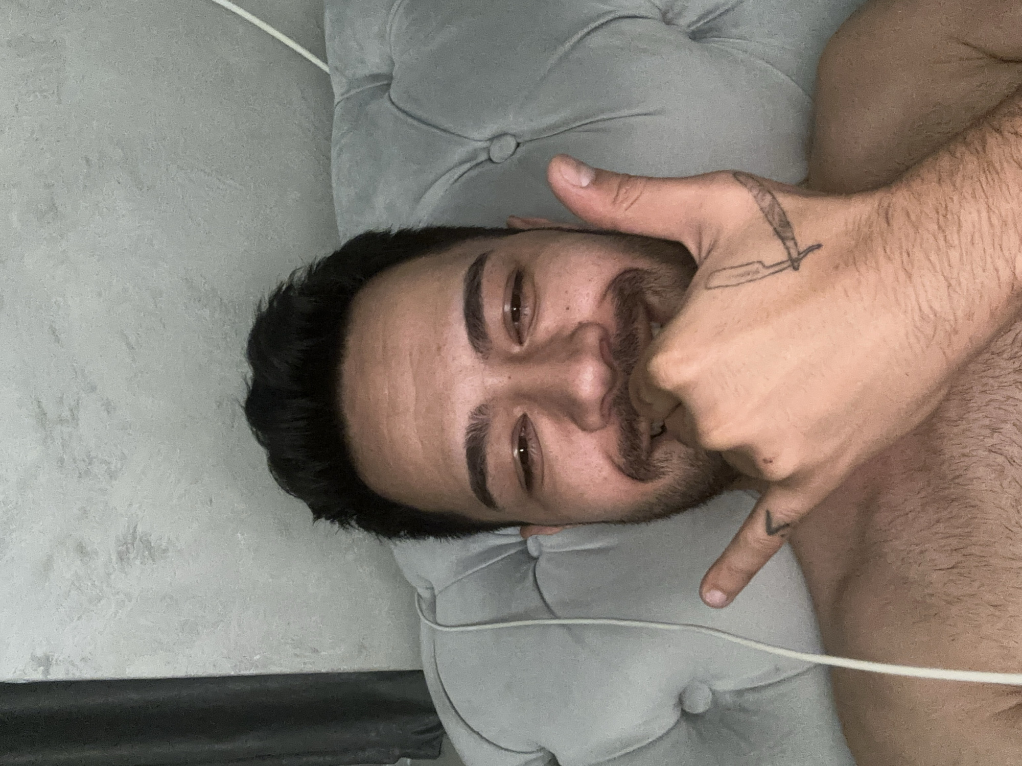 The image shows a man lying down indoors, positioned with his head towards the left side of the frame and oriented sideways. He rests his head on a light gray, tufted cushion or pillow. The man has dark hair, a mustache, and a short beard, and looks directly at the viewer with a relaxed, slightly smiling expression. His right hand is raised to his face, making a "shaka" or "hang loose" gesture, with his thumb and pinky extended and his middle fingers curled, covering part of his mouth.
Visible details on his body include a distinctive tattoo of a straight razor on the back of his right hand and a small "V" shaped tattoo on his right pinky finger. His bare shoulder and upper chest are visible, suggesting he might be shirtless. The background features a light gray, subtly textured wall or surface, with a thin white wire running diagonally across the upper portion of the image. A dark horizontal band, possibly part of furniture trim or a wall base, is visible at the very bottom.
The setting appears to be a cozy indoor environment, likely a home in Sofia, Bulgaria, given the provided context. The lighting suggests ambient indoor conditions. The overall mood is casual and comfortable, with the man appearing at ease. No discernible alphanumeric text is visible other than the symbolic tattoos.