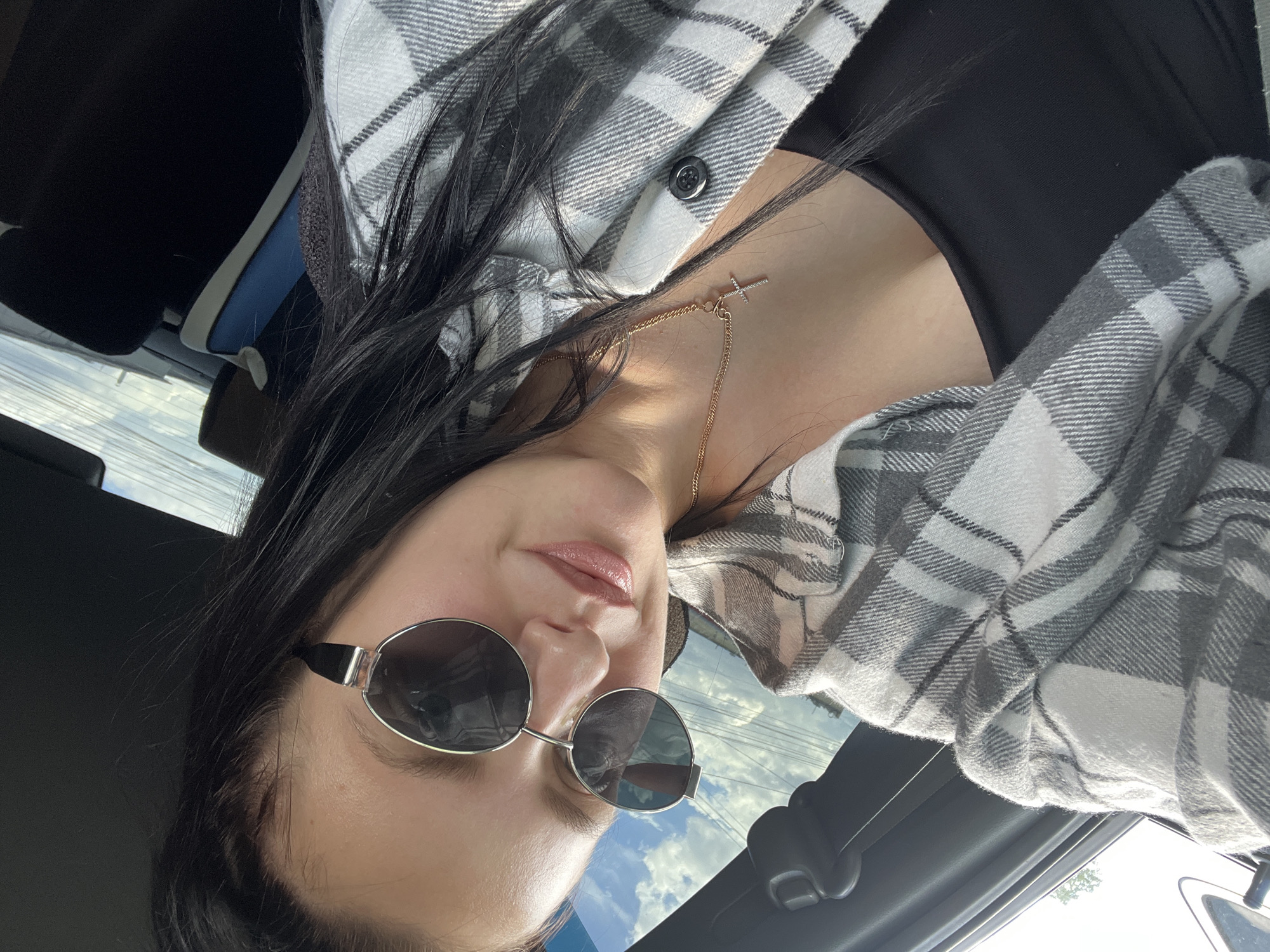 The image shows a person wearing sunglasses, a black top, and a grey and white plaid shirt, likely seated in a car. The reflection in the sunglasses suggests an outdoor environment, possibly a city street, with buildings and the sky visible. A gold necklace with a cross pendant is also visible around the person's neck. Considering the prompt, the location may be Chaikovsky, Russia, with the view from the car possibly including typical urban elements of that city.