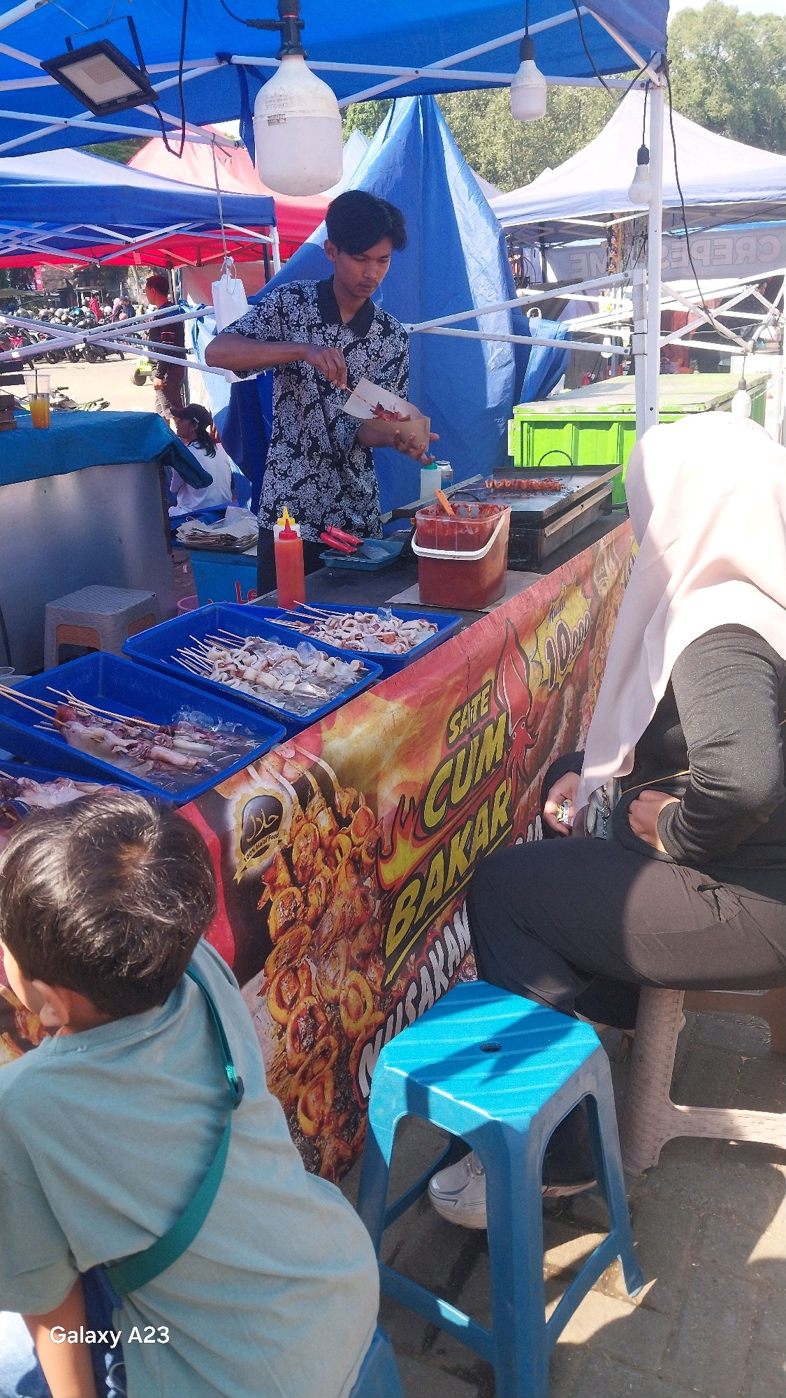 This image captures an outdoor food stall scene, likely in the city of Malang, Indonesia, given the visual cues and the "Galaxy A23" watermark. The stall is set up under a blue awning, with a red and yellow banner displaying text that appears to be in Indonesian, possibly advertising "Sate Cumi Bakar" (grilled squid satay).
A young man, dressed in a black and white patterned shirt, is actively preparing food. He is holding a piece of paper with grilled skewers on it and appears to be serving or arranging them. In front of him, a flat-top grill is visible, with some skewers already cooking. Several blue plastic trays containing raw squid on skewers are arranged on the counter. A red squeeze bottle, likely for sauce, and a large red container of sauce are also present.
To the right of the stall, a woman wearing a light-colored hijab and a black outfit is seated on a chair, facing away from the camera. Her hands are resting on her lap. In the foreground, the back of a young boy wearing a light blue t-shirt with a green strap over his shoulder is visible. He is seated, facing the food stall. Another blue plastic stool is positioned in front of the woman. The ground appears to be paved. In the background, other tents and stalls are visible, suggesting a market or a street food gathering. The overall lighting suggests it is daytime, with sunlight casting shadows.