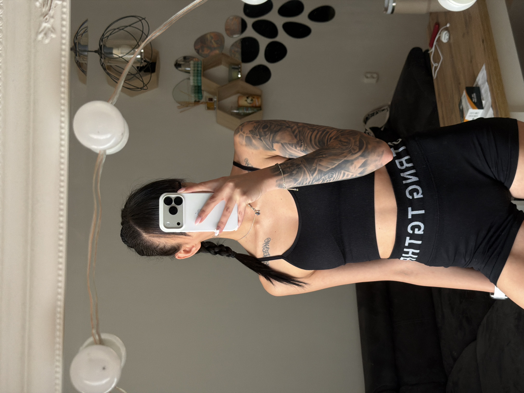 This is a vertical, medium shot photograph of a person taking a selfie in a mirror. The person has dark hair styled in a braid, and is wearing a black tank top and black athletic shorts with white lettering on the waistband. They have extensive tattoos on their left arm and a small tattoo on their chest. A silver necklace with a blue pendant is visible. The person is holding a white smartphone to take the picture.
The background reflected in the mirror shows a room with modern decor. Visible objects include two white pendant lights hanging from the ceiling, a decorative wire lampshade, and shelves on the wall holding various items. There are also decorative, irregularly shaped mirrors on the wall. A wooden table with some items on it, including what appears to be packaging for a product, is also visible. A dark sofa is partially visible in the lower right corner of the image. The lighting suggests it is daytime.
The white lettering on the shorts reads "FITNESS".
The context indicates the location is Plovdiv, Bulgaria. No specific emotions are conveyed, and the activity is simply taking a selfie.