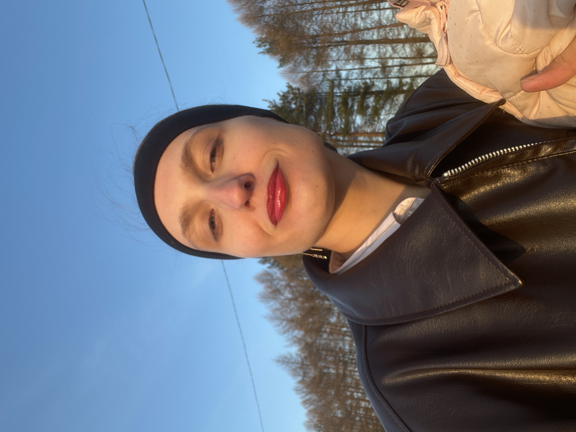 A woman with pale skin, wearing a black headband and red lipstick, is seen in a low-angle selfie outdoors. She is wearing a dark brown leather jacket over a white shirt. The background consists of a clear blue sky, bare trees, and what appear to be power lines. The location is potentially Подшивалово, Russia, based on the user's request.