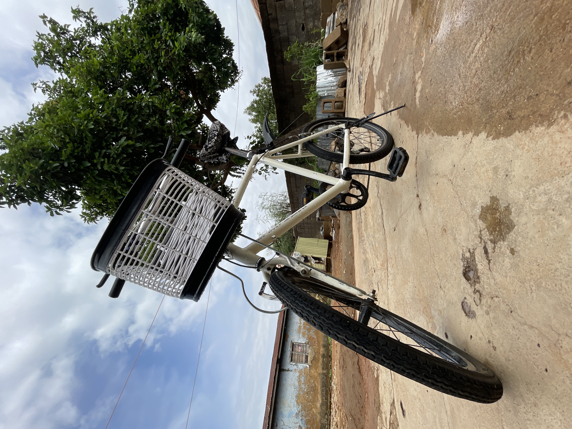 A low-angle shot captures a white bicycle with a black and white woven basket on its front handlebars, parked outdoors. The bicycle's frame bears partially visible text that reads "all good things with good pe." Its front wheel, with a textured tire, is prominently featured in the foreground, while the rear wheel and pedal are also visible.
To the left and above the bicycle, a large, verdant tree with dense green foliage fills a significant portion of the frame, set against a bright, partly cloudy blue sky.
To the right and below the bicycle, the ground is a rough, textured concrete or paved surface, showing stains and potentially wet areas. In the mid-ground, behind the bicycle, several simple, weathered buildings with corrugated metal roofs and rough walls, possibly concrete or brick, are visible. Scattered nearby are loose construction materials, including cinder blocks and corrugated metal sheets.
The scene depicts a quiet daytime setting in what appears to be a semi-urban or rural environment in Jos, Nigeria, characterized by modest architecture and natural elements. No individuals are present in the image.