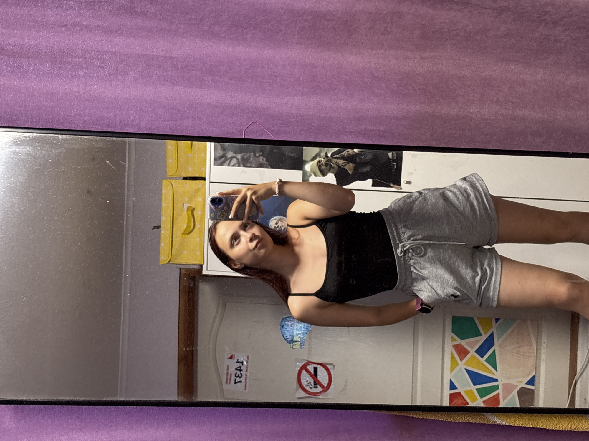 A young woman with long, dark hair is taking a mirror selfie in what appears to be a bedroom. She is wearing a black tank top and grey sweat shorts. The mirror reflects a cluttered room with a purple curtain on the right side. Behind her, on the left, is a white door with various stickers and a colorful geometric art piece. On top of the door are two yellow boxes. The mirror itself is dusty and shows reflections of posters on the wall and the ceiling. The overall impression is a casual, personal space. The city of Прикамский, Russia, is not identifiable from the visual cues in the image.