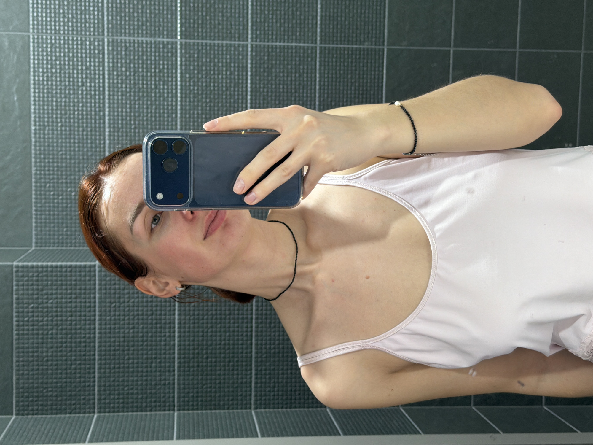 This image depicts a female individual taking a mirror selfie in an indoor setting within the city of Chaikovsky, Russia. The subject, with reddish-brown hair pulled back, is visible from the upper torso upwards. She is wearing a light pink camisole, a thin black choker necklace, and a black beaded bracelet with a single white pearl on her left wrist. A small, silver-colored hoop earring is visible on her right ear. Her left hand holds a dark blue smartphone, partially obscuring her face below the eyes. The smartphone, enclosed in a clear case, shows a triple-lens camera array on its rear. Her expression features a slight smile. The background consists of dark grey rectangular tiles with a raised, textured grid pattern, forming a wall. A lighter, vertical strip is visible along the left edge of the frame where the tiled wall meets another surface. The lighting is uniform, suggesting an interior space.