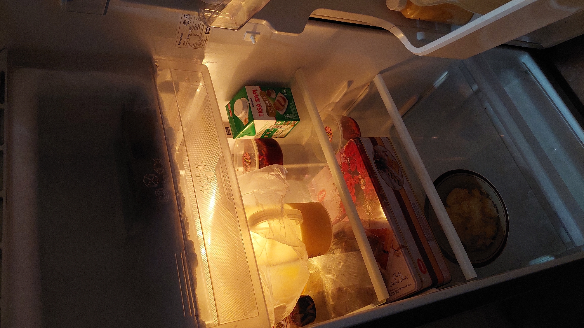 This is a forensic, matter-of-fact description of the inside of a refrigerator. The refrigerator is illuminated by its internal light, casting a warm glow on the contents. On the top shelf, there are two clear plastic containers, one filled with a dark red substance, possibly jam or preserves, and another larger container covered in plastic wrap containing a yellow liquid, likely juice or a beverage. To the right of these is a green and white carton of "Tiga Sapi Evaporasi" milk, a product commonly found in Indonesia.
Below this shelf, on the middle shelf, is a long, rectangular box with an image of red roses on it, possibly containing food items. To the left of this box, partially obscured, are more clear plastic containers with red contents.
The bottom shelf contains a dark plate with a mound of yellowish, granular material, which could be cooked rice or a similar food item. The interior walls of the refrigerator show signs of condensation or frost build-up, particularly near the top. A label on the interior right wall of the refrigerator shows "Panasonic" and other product information, including a model number and manufacturing details for Indonesia.
The location is identified as Pulongrambe, Indonesia, suggesting this is a domestic refrigerator within a residence in that area. No people are present in the image, and no actions or interactions are depicted. The environment is solely the interior of the refrigerator.