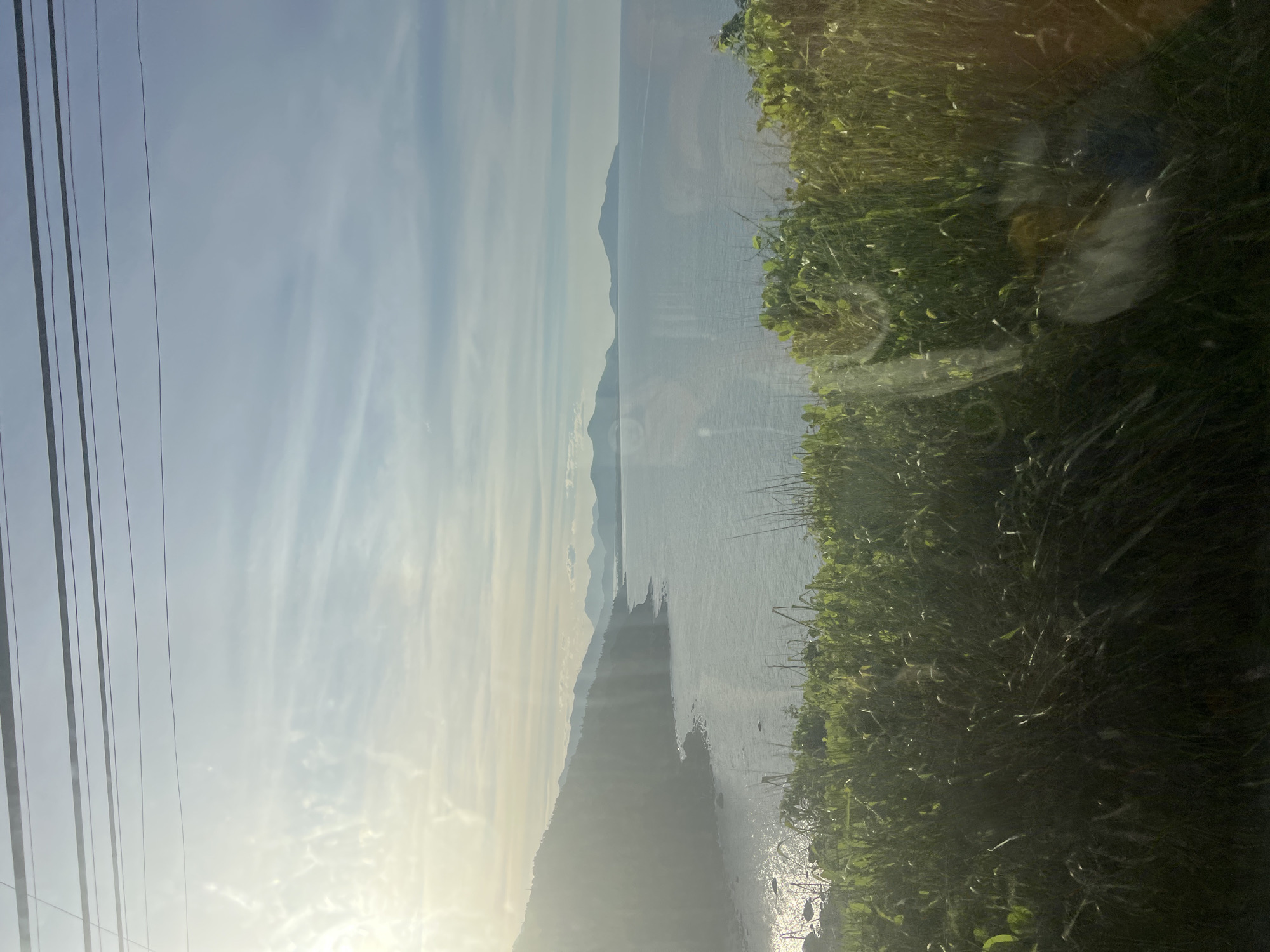 This is a landscape photograph taken from an elevated vantage point, likely from a moving vehicle, overlooking a coastal scene. In the foreground, dense, green grass and foliage are visible, creating a natural border. Beyond the vegetation, a calm, blue sea stretches out, meeting a distant shoreline. A chain of mountains, some appearing as islands, are silhouetted against the horizon under a pale blue sky with wispy clouds. Sunlight streams in from the left, casting a bright glow on the water and the distant mountains, creating a hazy effect. Several dark, horizontal lines, likely power lines, are present across the top of the frame. The image conveys a serene, natural environment, characteristic of the coastal areas near Huế, Vietnam, known for its picturesque seascapes and mountainous terrain.
