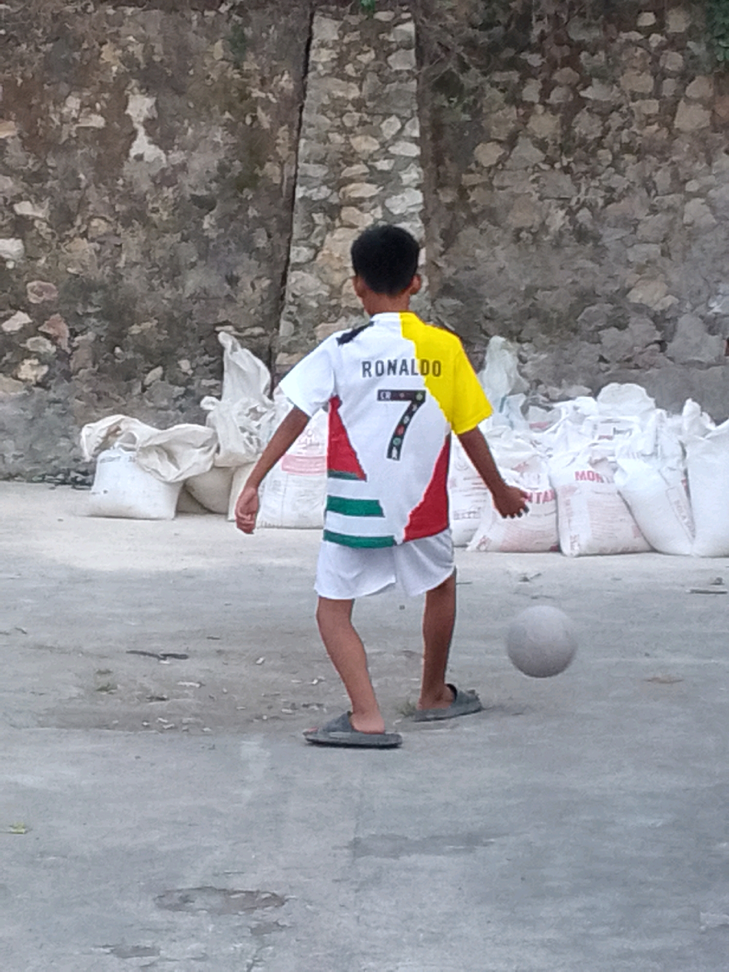 An outdoor, eye-level shot captures a young boy in the city of Gunungmasigit, Indonesia, engrossed in playing soccer. The boy, seen from behind, wears a white shirt with yellow sleeves, emblazoned with "RONALDO" and the number "7" in black. He also sports white shorts and grey flip-flops. His left arm is extended, suggesting he is in the process of kicking or controlling a light gray soccer ball, which hovers a few inches above the concrete ground. The background features a textured stone wall and several large, white sacks that appear to be stacked. The ground is a rough, weathered concrete surface with scattered debris.