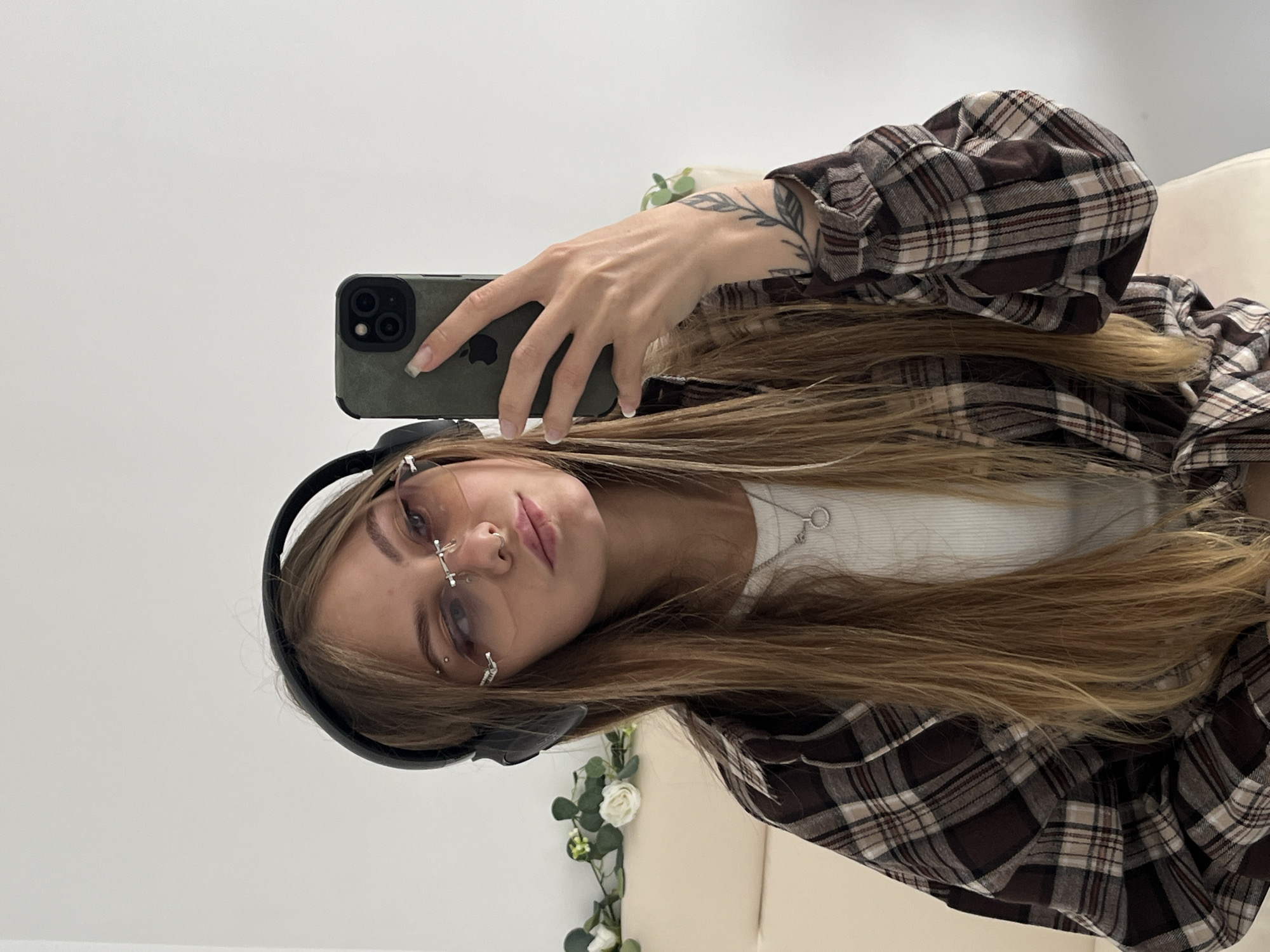 A young woman is taking a selfie with a phone in a green case. She is wearing headphones, glasses with a nose ring attachment, a white shirt, and a brown plaid flannel shirt. Her blonde hair is long and flowing. She has a tattoo on her inner forearm. Artificial flowers are visible in the background. The environment appears to be indoors with white walls. The location is unknown and there is no information on the city.