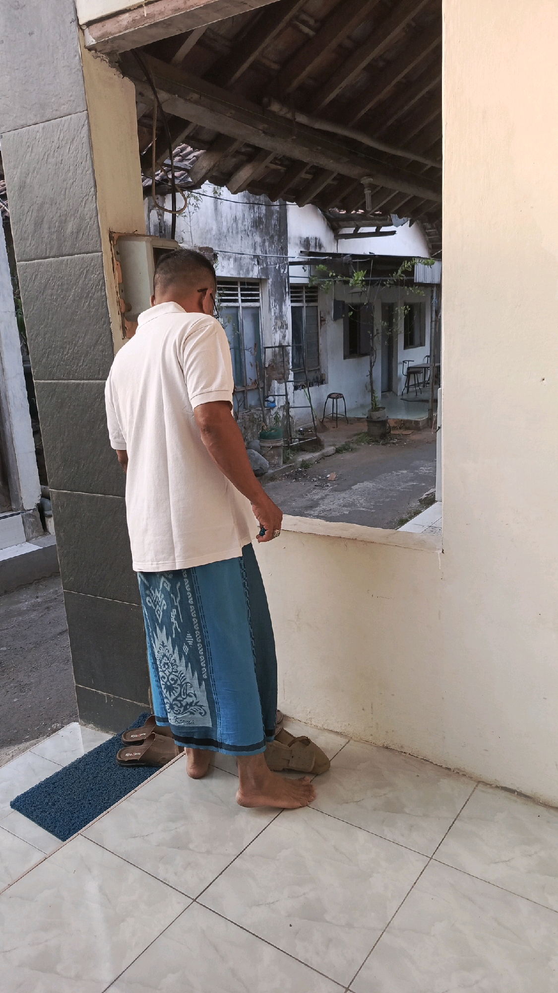 A man stands facing away from the camera, looking out of a wide window. He is wearing a light-colored polo shirt and a blue sarong with a patterned design. His feet are bare, and he stands on a tiled floor. To his left, on a dark blue mat, are two brown sandals. To his right, another pair of brown sandals rests on the tile. The scene appears to be an outdoor or semi-outdoor area of a building, possibly a house or a small commercial establishment. The background shows a worn, weathered building with visible structural elements and some vegetation. The image is taken in Pekauman Kulon, Indonesia.