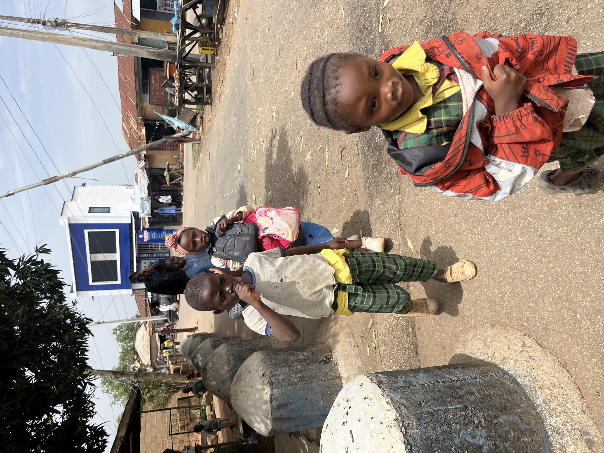 Three children stand on a paved street in Jos, Nigeria. The child closest to the viewer wears a yellow shirt, green plaid pants, and a red jacket, and faces the camera. Behind them is a child wearing a pink head covering and a child wearing a white shirt and green plaid pants. The children are standing next to a series of gray, rounded concrete posts. In the background, a utility pole, buildings with corrugated metal roofs, and a building with a blue facade can be seen. Other people are visible in the distance.