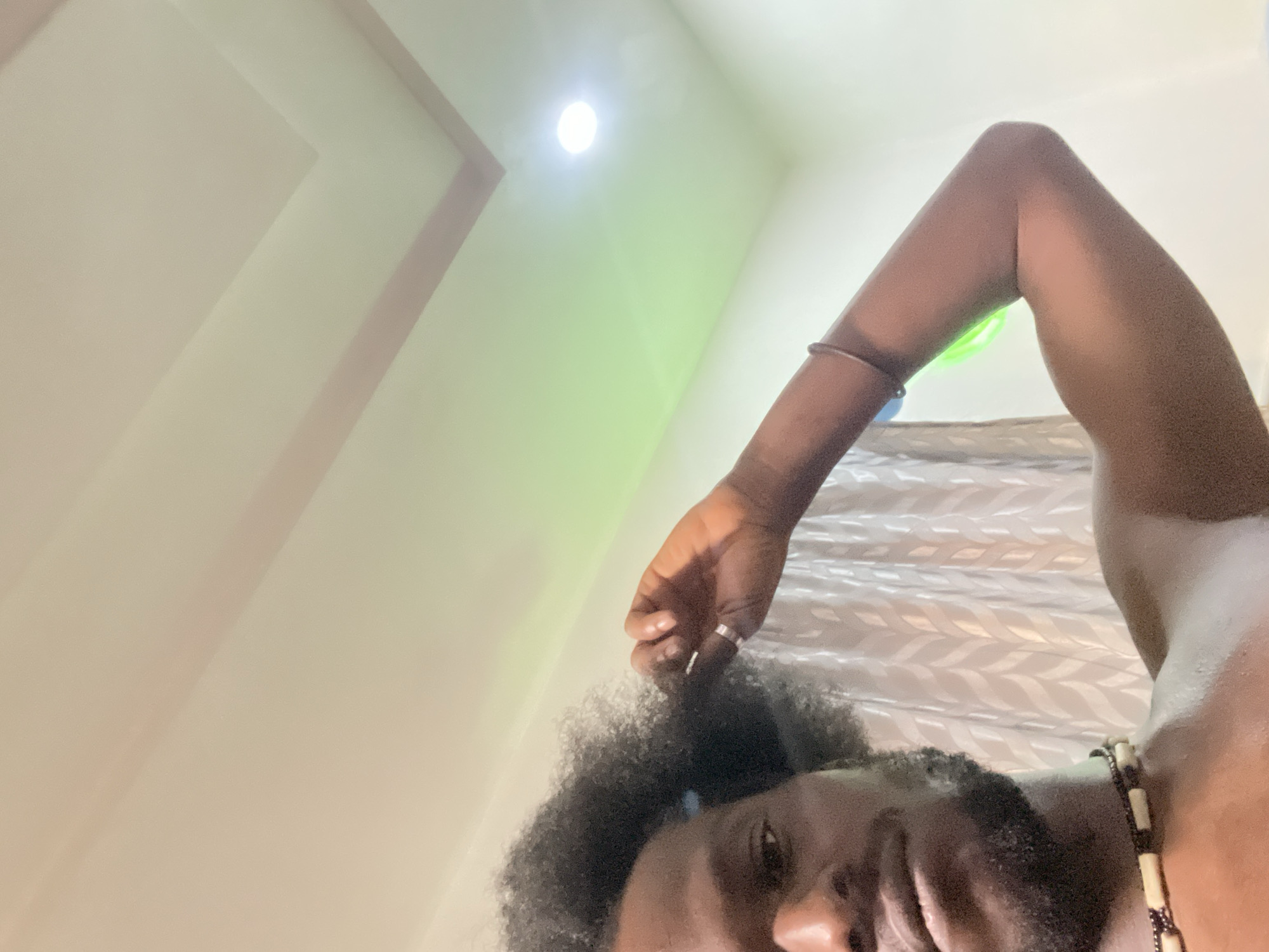 A low-angle shot taken indoors reveals a person of African descent in Kuchingoro, Nigeria. The individual has dark skin, an afro hairstyle, and a beard. The person's right arm is raised, with a silver ring visible on the ring finger. A silver bracelet is worn on the forearm. A necklace with light-colored beads is around the neck. The background shows a white wall with a light fixture. A bed with a patterned white bedsheet is partially visible.