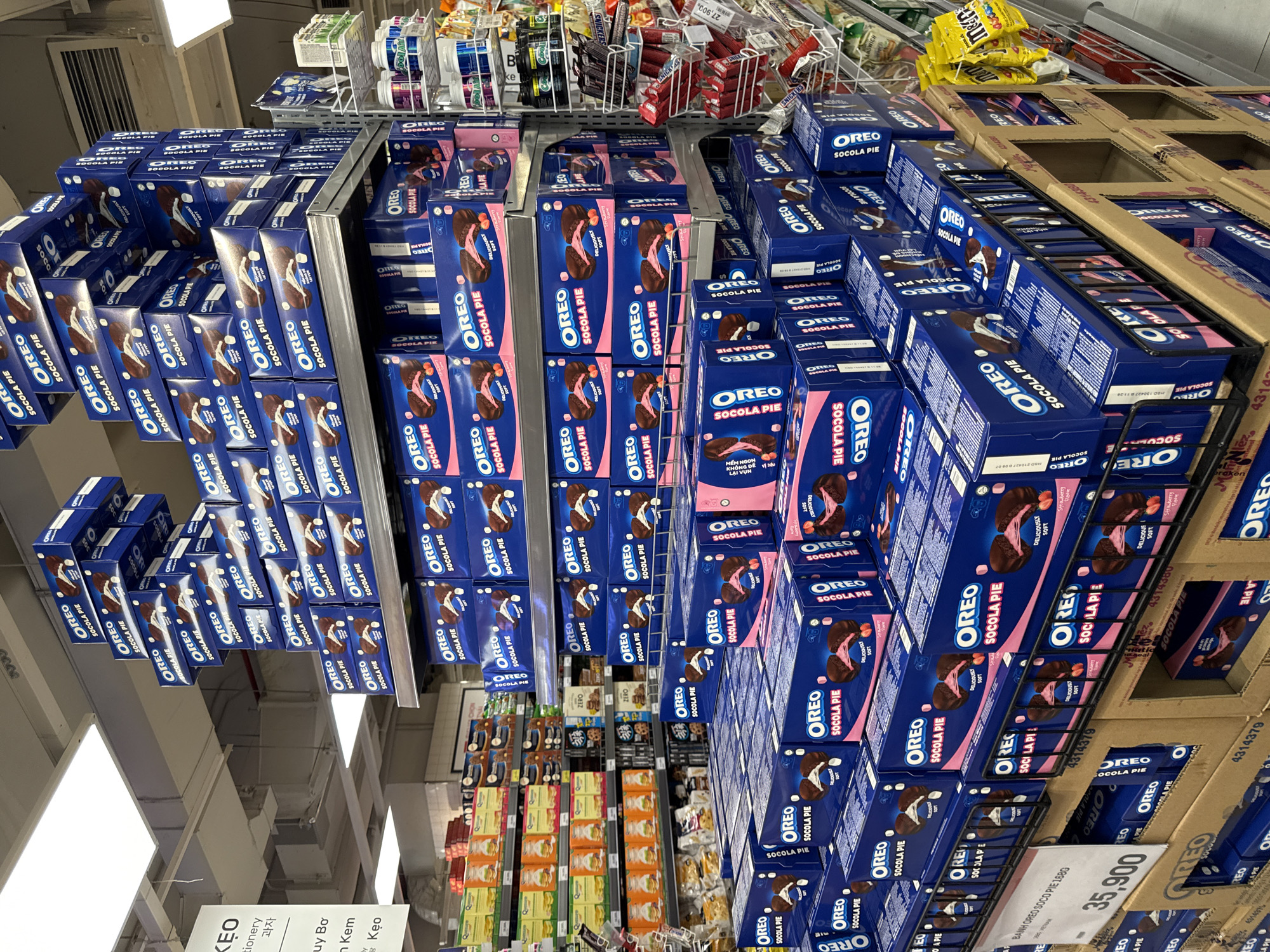 The image shows a supermarket aisle in Nha Trang, Vietnam, featuring a large display of Oreo "Socolia Pie" products. The products are stacked high on shelves, with multiple boxes visible. The boxes are predominantly blue with pink accents and feature images of the cookie. The lighting suggests an indoor retail environment. On the left side of the image, a sign with Vietnamese text is partially visible. The overall impression is of a well-stocked confectionery aisle in a supermarket.