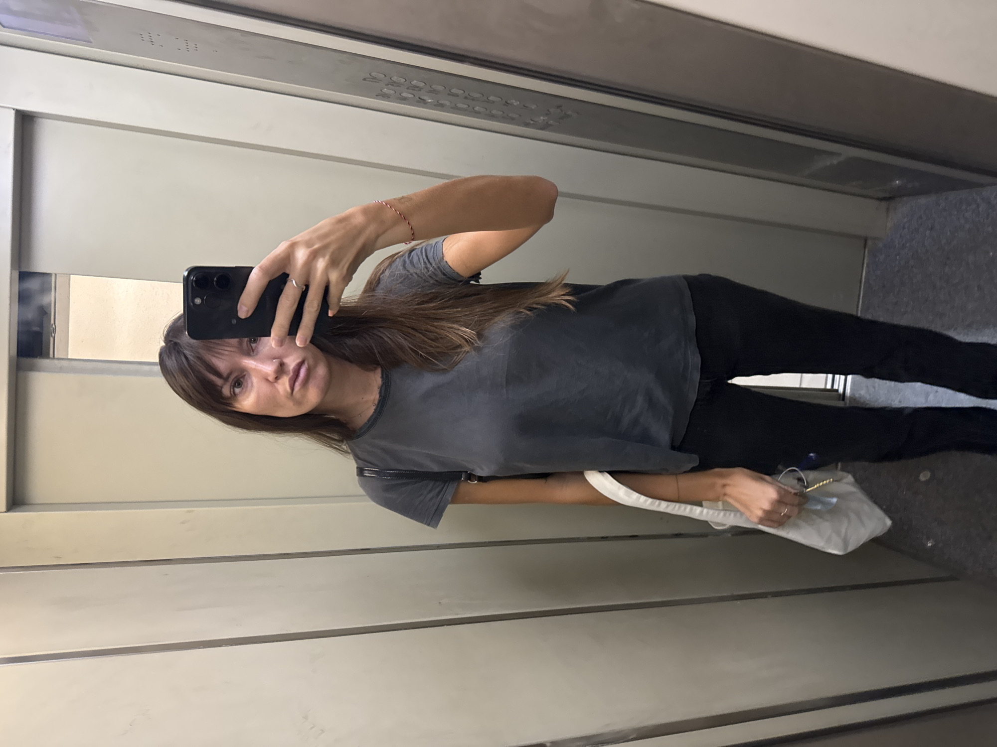 In an elevator in Moscow, Russia, a woman with long, dark hair and bangs stands facing a phone held for a selfie. She wears a dark grey t-shirt and black pants. A ring is visible on her left ring finger. She carries a white tote bag over her left shoulder, holding keys and a cup in her hand. The elevator walls are metallic grey, with vertical seams. The floor is speckled grey. The elevator door is partially open, revealing a glimpse of the adjacent space.