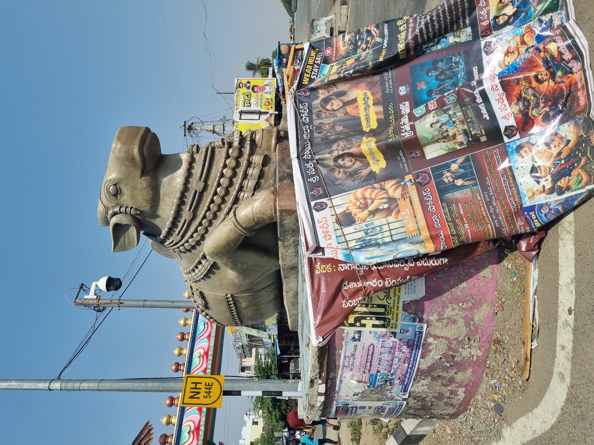 The image shows a stone statue of a bull adorned with beaded necklaces, located in Kodikonda Check Post, India. The statue is next to a road and has a column covered with numerous posters and signs. A yellow road sign marked "NH 544E" is visible on a pole near the statue. One of the prominent posters encourages wearing a helmet and staying safe. Other posters contain safety messages from the Satya Sai District Police, in Telugu. A security camera is mounted on a nearby pole. There are a few people visible near the base of the statue. Buildings and power lines are visible in the background under a clear blue sky. The overall environment is a busy roadside with a blend of religious imagery, safety messages, and commercial advertising.