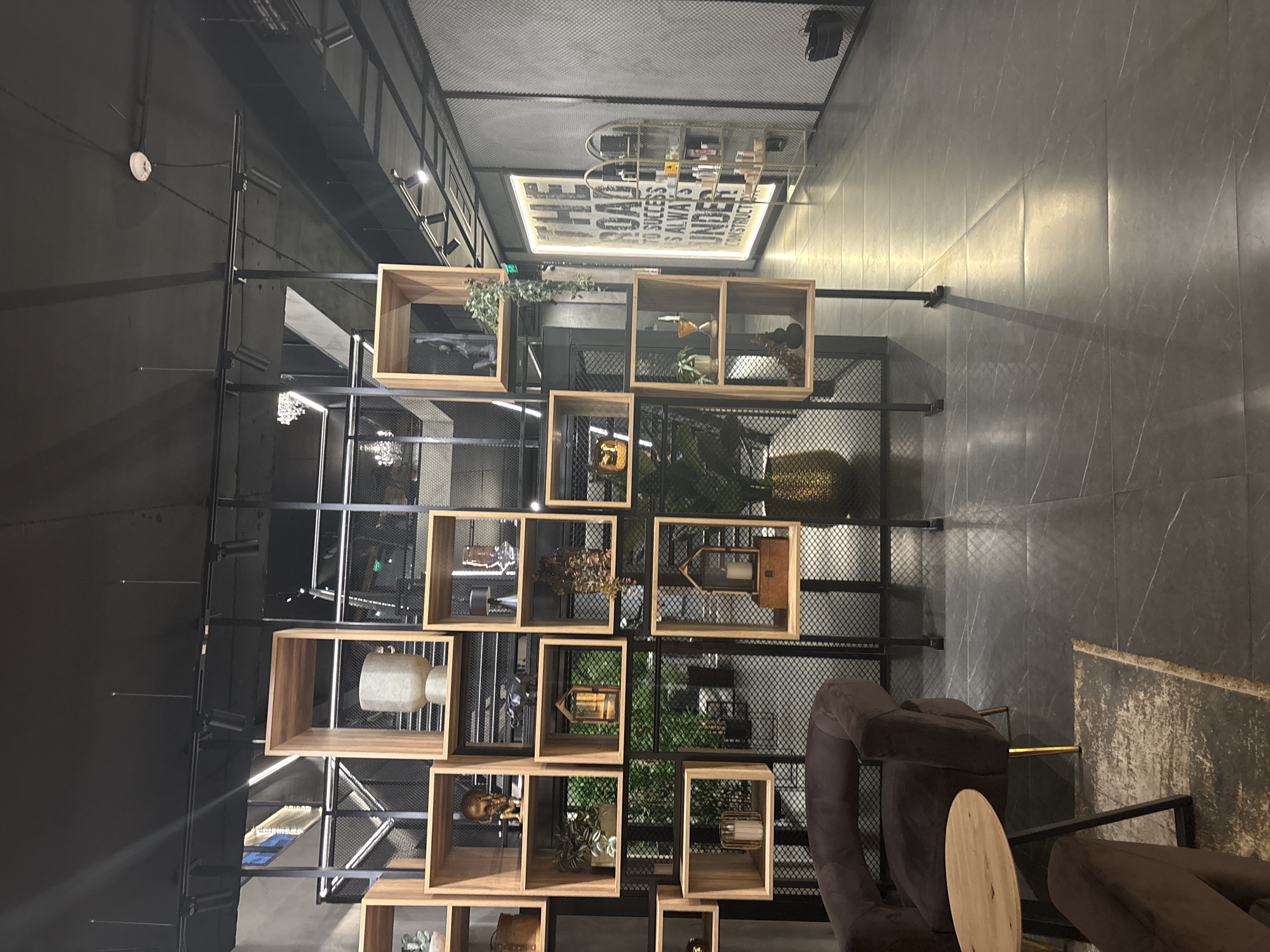 This image shows an indoor space in what appears to be a cafe or lounge with modern, industrial-chic decor. The dominant feature is a large, black metal shelving unit filled with various decorative objects, including plants, vases, and small sculptures. The shelving unit is composed of open wooden cubes and rectangles of varying sizes. The background behind the shelving appears to be a dark grey mesh.
The foreground includes comfortable-looking brown armchairs with a small wooden table between them. The lighting is somewhat dim, creating a relaxed ambiance.
The location is unidentified, and it's not confirmed if it's located in Plovdiv, Bulgaria. There are no people visible in the image.