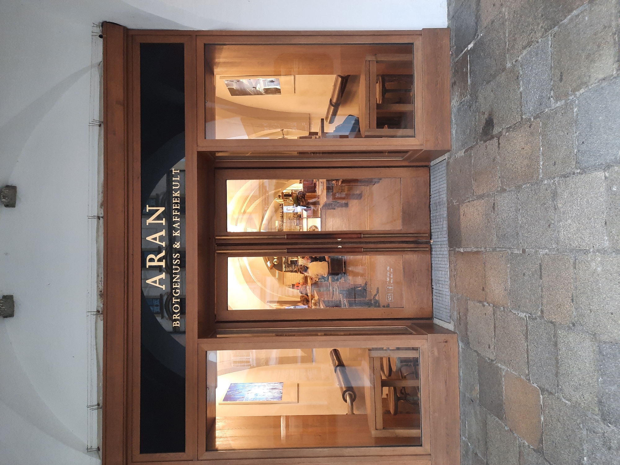 The image shows the exterior of a shop called "ARAN BROTGENUSS & KAFFEEKULT" in Rosenheim, Germany. The shop has a wooden facade with large glass windows and double doors. Inside, several people are visible sitting at tables, suggesting it is a cafe or bakery. The storefront is decorated with framed pictures and displays of baked goods or bread-making equipment. The pavement outside is made of stone. The overall scene depicts a commercial establishment in an urban setting.