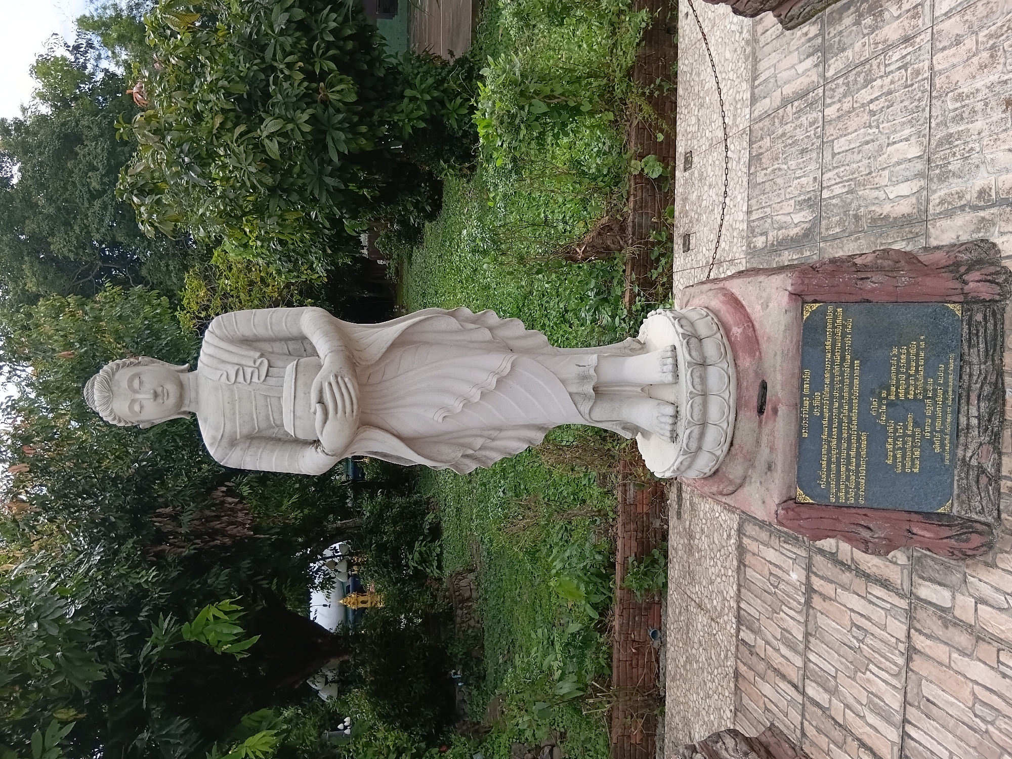 A full-shot photograph captures a serene white stone statue of Buddha standing in a garden setting. The Buddha is depicted in a calm, upright posture, holding a bowl in his clasped hands. He is dressed in traditional monastic robes that fall gracefully around his form. The statue stands on a tiered, ornate base, which is itself mounted on a pedestal. This pedestal features a dark, rectangular plaque inscribed with Thai script, likely detailing the statue's history or significance. The base of the pedestal is textured and appears to be made of a reddish-brown material, possibly stone or concrete, resembling rock formations.
The statue is situated outdoors, with lush green foliage and trees forming the backdrop. The ground in front of the pedestal is paved with light-colored rectangular tiles arranged in a pattern. A low brick wall runs along the edge of the paved area, separating it from the more overgrown garden beyond. The garden itself is densely planted with various types of vegetation, creating a natural and somewhat wild environment. In the background, glimpses of other structures and elements of the garden, such as what appear to be decorative lamps and possibly a golden structure, are visible through the trees. The lighting suggests it is daytime, with diffused light indicating overcast conditions or dappled shade from the trees. The image is taken from a slightly low angle, emphasizing the height and presence of the Buddha statue. The location is identified as Ban Ruammit, Thailand.