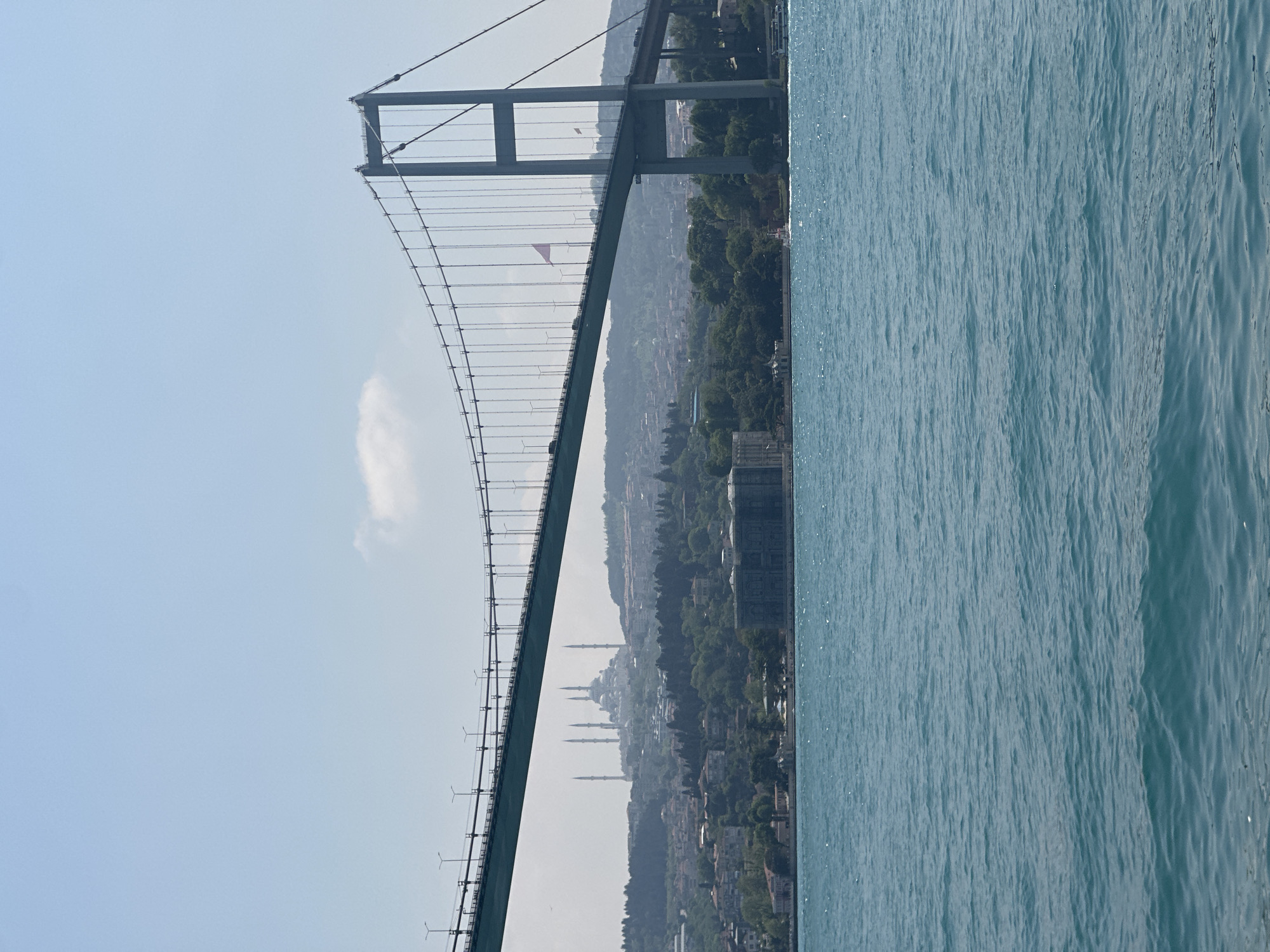 The image depicts the Bosphorus Bridge in Istanbul, Turkey, connecting Europe and Asia. The bridge spans a body of water. On the Asian side, the skyline is visible, revealing minarets, suggesting the presence of mosques in the city. The bridge itself is a suspension bridge with vertical suspension cables supporting the deck. The sky is overcast with a light haze. The body of water occupies the right side of the frame, featuring gentle waves. The scene appears to be captured during daylight. The city is in Beşiktaş, Turkey.