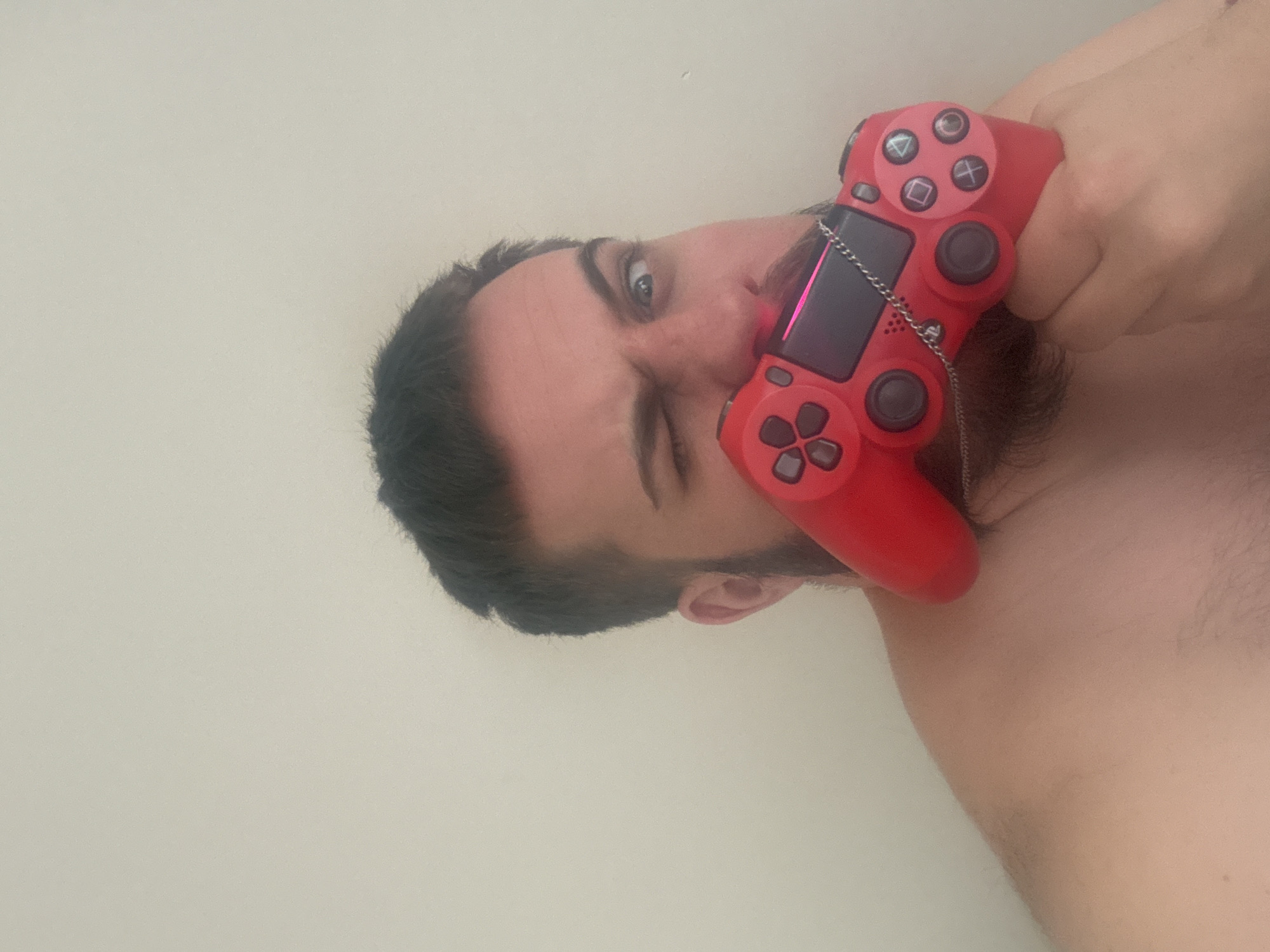 A shirtless man is holding a red PlayStation 4 controller to his face, winking with his left eye. He has dark hair and a beard. The background is a plain, light-colored wall. There is no indication of location or city.