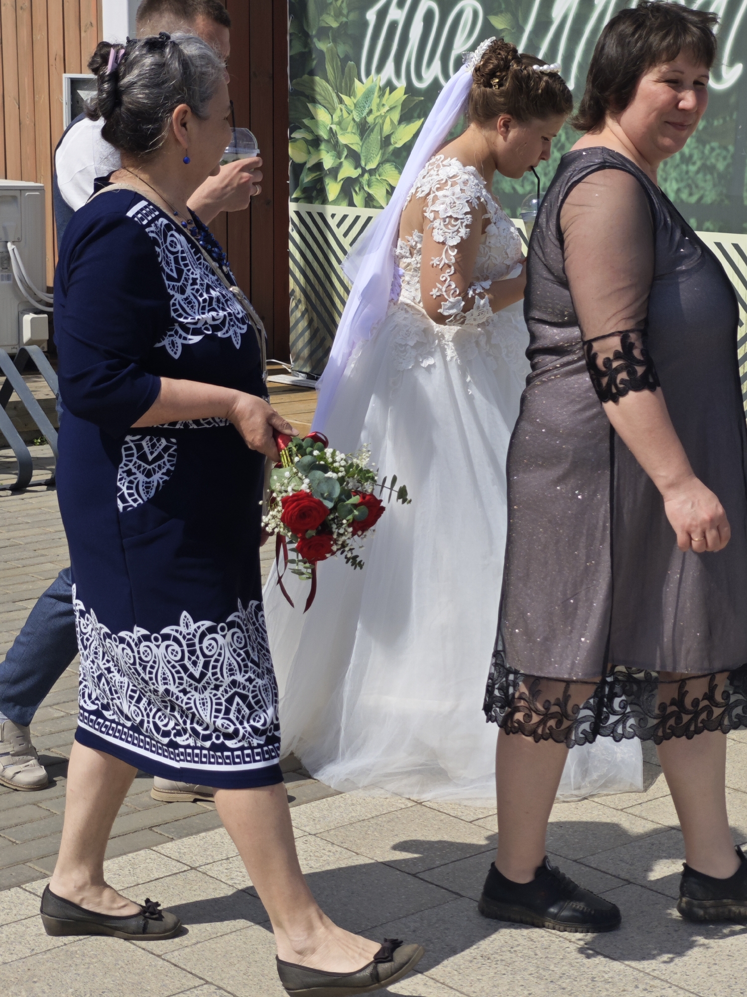 In Chaikovsky, Russia, a bride in a white wedding dress with lace detailing and a long veil walks on a paved surface, likely a sidewalk. She appears to be drinking from a clear plastic cup with a straw. To her right is a woman in a knee-length gray dress with sheer sleeves and ornate black lace trim at the cuffs and hem. To the bride's left, a woman in a navy knee-length dress with white lace patterns holds a bouquet of red roses. A man in jeans and a white shirt walks behind the first woman, holding a clear cup. Another man is partially visible at the top of the frame. A building with a striped wall and green foliage is in the background.