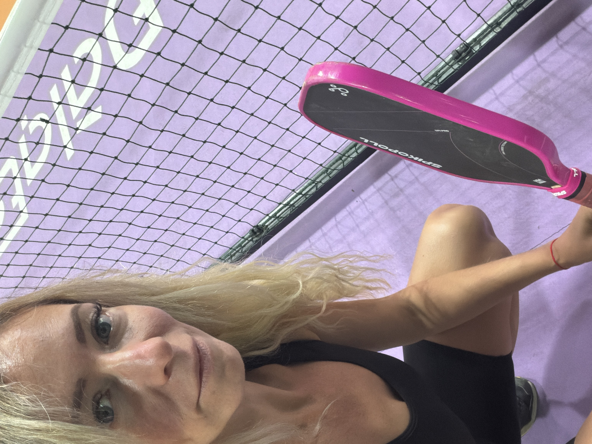 A blonde woman wearing a black athletic outfit squats in front of a net, holding a pink and black paddle. The background is a pale purple surface with a diamond-patterned net. The woman's face is partially visible, with blue eyes and blonde hair. The paddle has text that reads "SPIKEPOLL". The scene appears to be an indoor court.