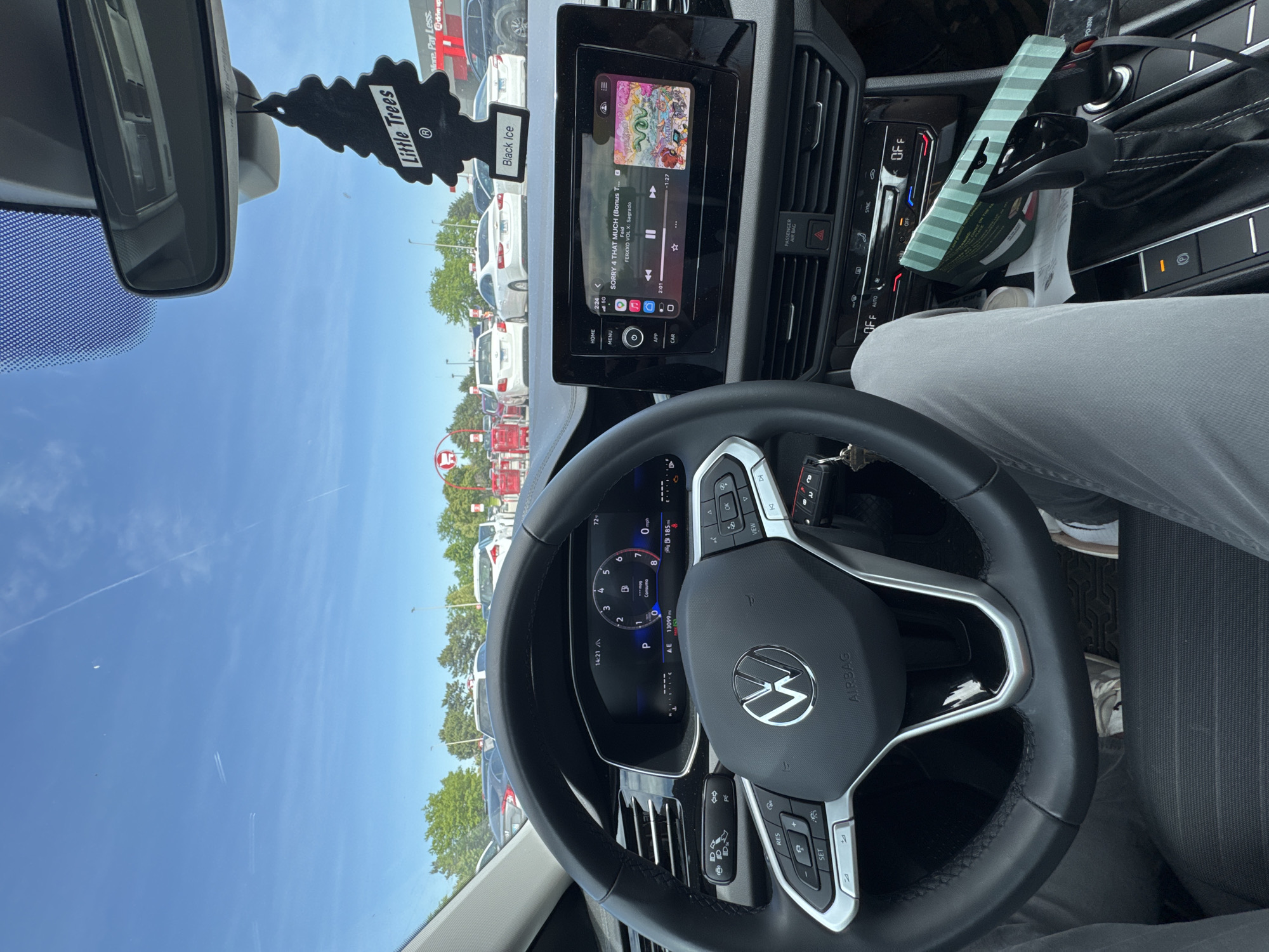 The image shows the interior of a car, likely a Volkswagen, with a clear view through the windshield of a Target store in Glendale Heights, Illinois, United States of America. The dashboard features an infotainment screen displaying music ("SORRY 4 THAT MUCH" by Feid) and climate controls. A "Little Trees" air freshener (Black Ice scent) hangs from the rearview mirror. The driver's hands are on the steering wheel, and the odometer reading is 13099 miles. A drive up order is visible with the words "More Pay Less" and "Drive upa" in the upper right corner.