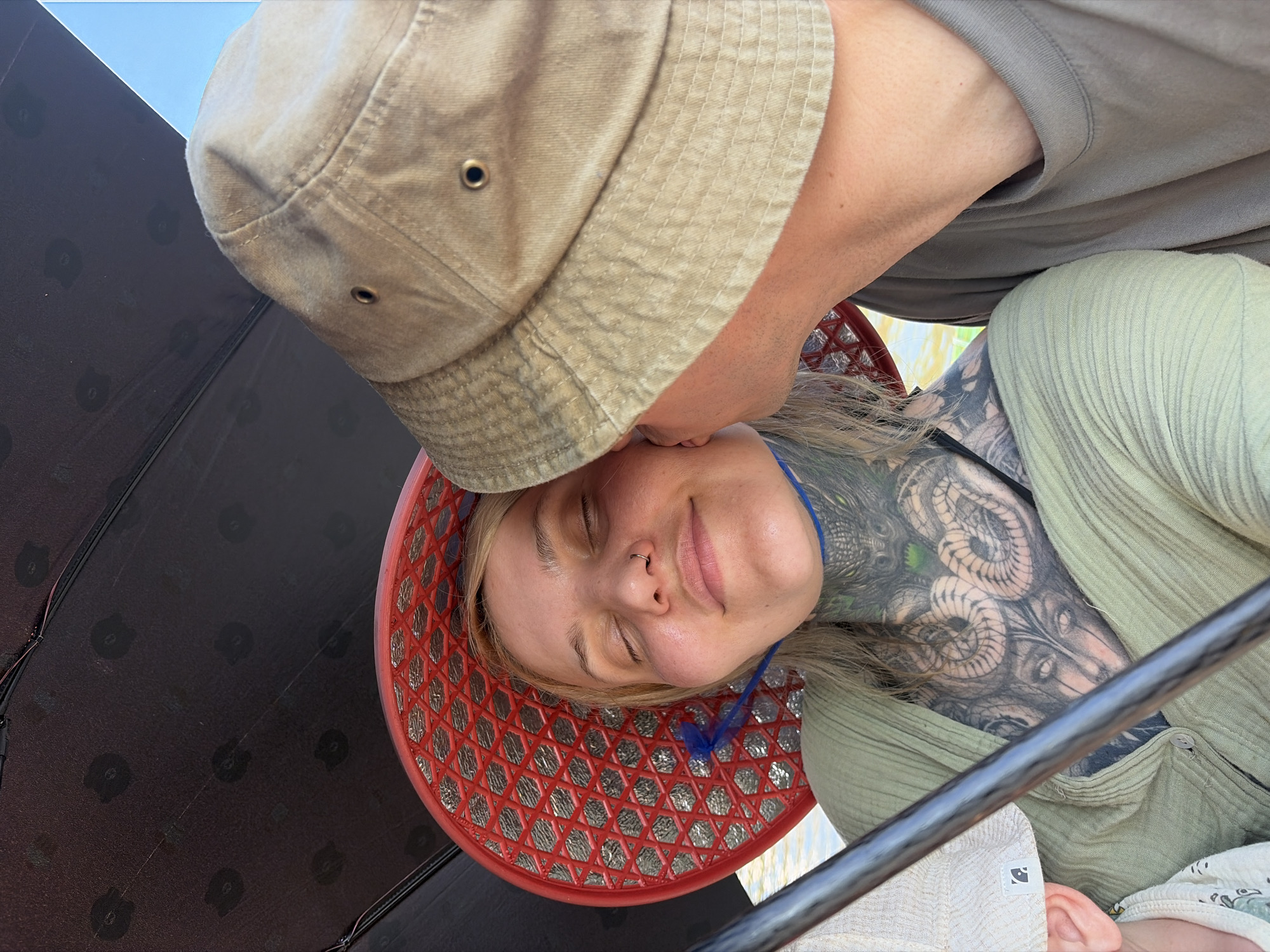 This image captures an intimate and affectionate moment between two people in an outdoor setting, likely in Đà Nẵng, Vietnam. One individual, wearing a beige bucket hat and a grey t-shirt, is seen leaning over to gently kiss or nuzzle the forehead of the second person. This person, who has blonde hair and a septum piercing, is lying down with her eyes closed, displaying a serene and contented smile. She is adorned with extensive, intricate tattoos covering her chest and left arm, featuring patterns that resemble coiled snakes or mythical creatures. She wears a light green, ribbed top, and a blue string is visible around her neck.
Her head rests on a distinctive red, woven object with a lattice pattern, possibly a hat or tray, which adds a cultural touch to the scene. The bright blue sky in the background indicates daytime, while a large dark umbrella or canopy on the left provides shade, suggesting a sunny environment. A metallic bar is visible diagonally across the lower right portion of the frame. The image beautifully conveys feelings of tenderness, relaxation, and mutual affection. No discernible text is visible within the frame.